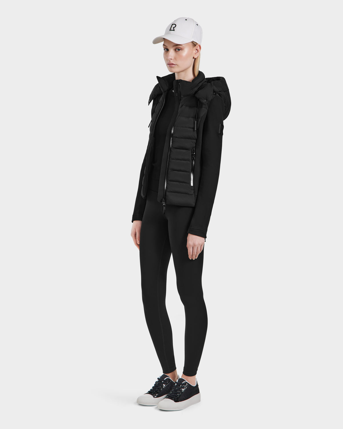A person faces slightly to the side, wearing a white cap and the Rudsak MABS - 8124825 Black womens hooded puffer vest over a black long-sleeve top with leggings and sneakers, set against a plain white background.