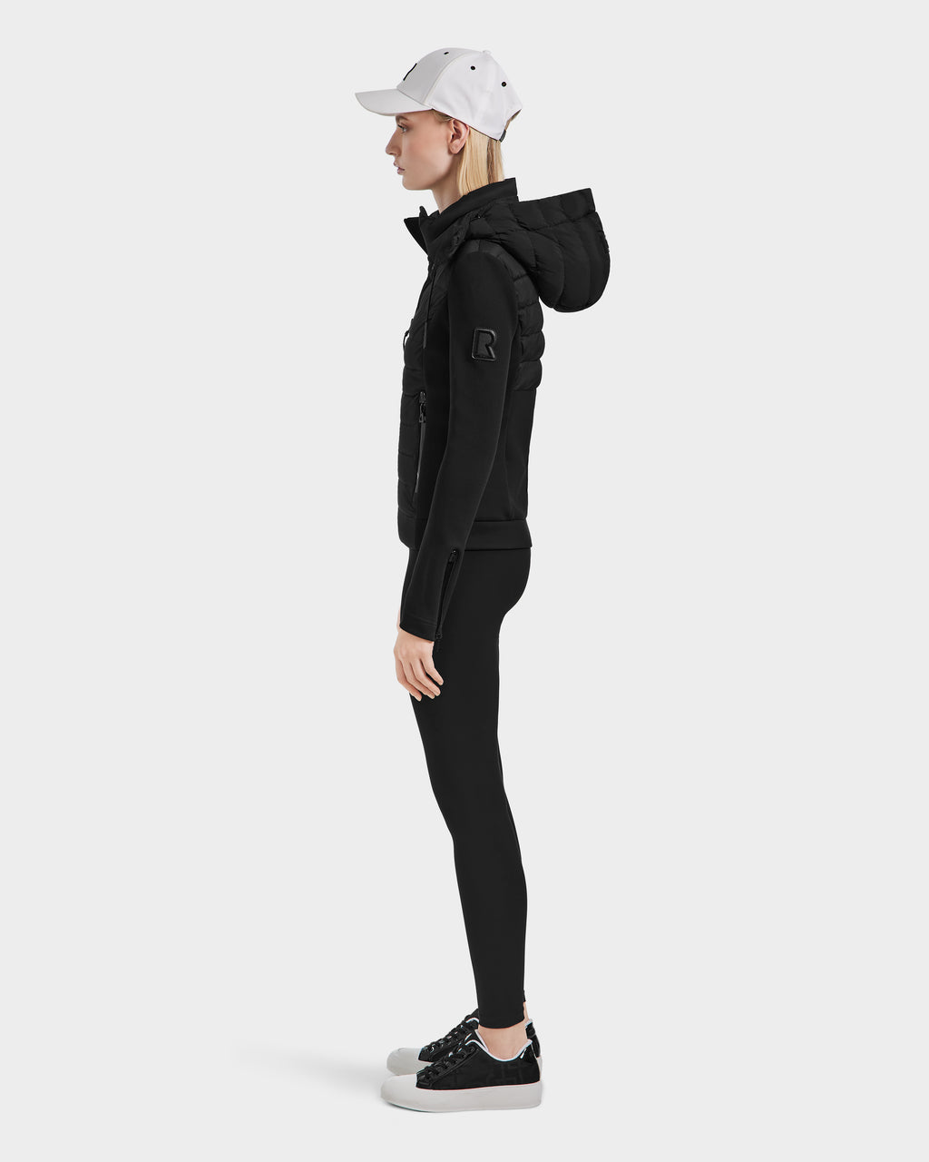 A woman in a white baseball cap and Rudsak MABS - 8124825 Black neoprene jacket, black leggings, and black sneakers stands in profile against a plain white background.