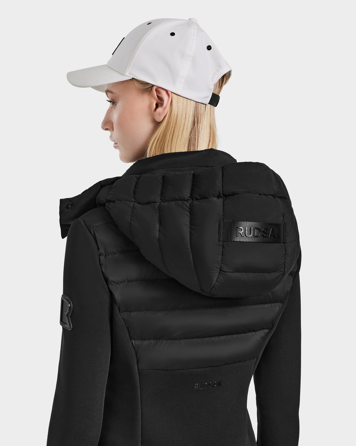 A woman in a white cap models the Rudsak MABS - 8124825 Black jacket, featuring quilted details and branded hood, shown from the side and back against a plain background.