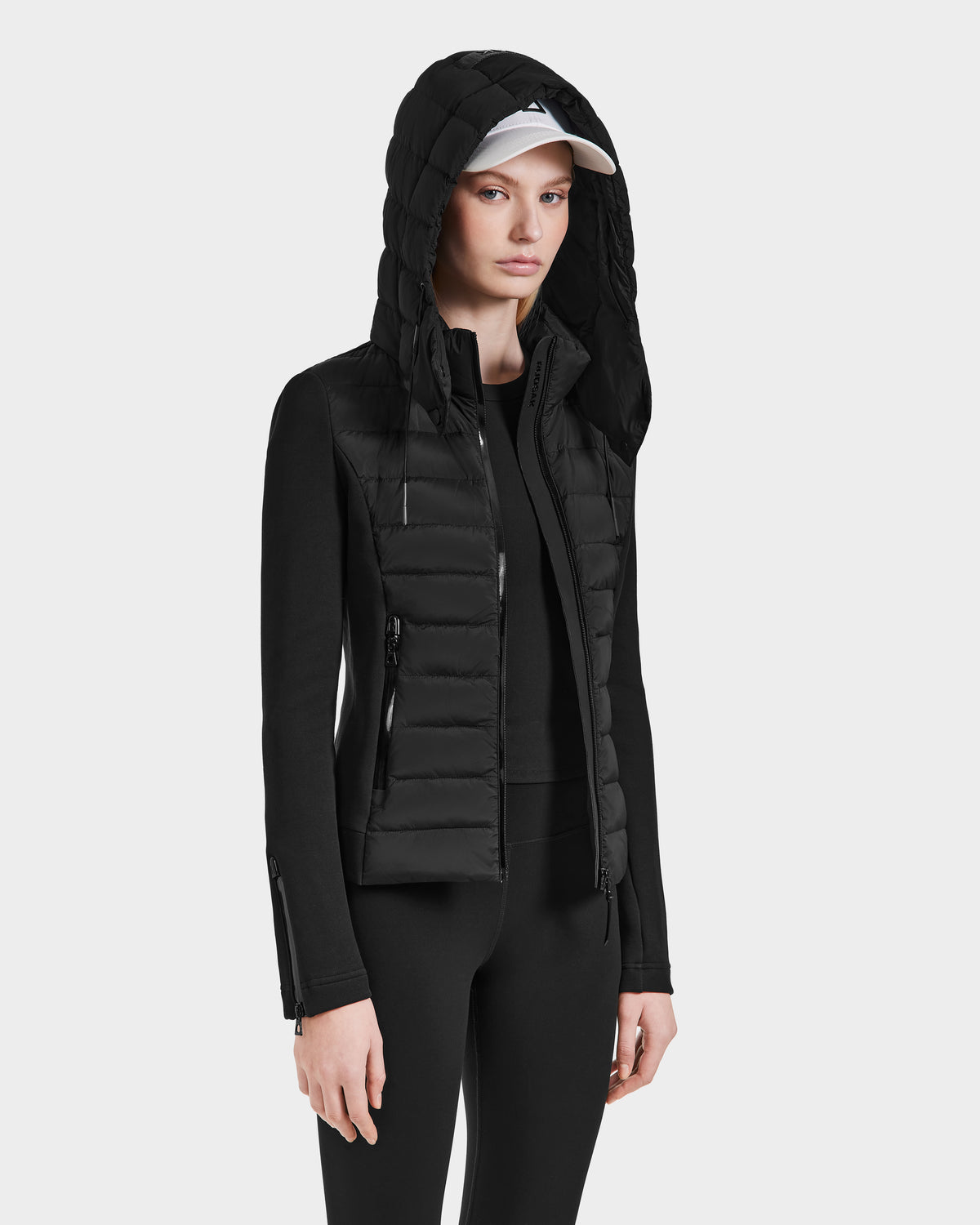 A woman wearing the Rudsak MABS - 8124825 Black lightweight hooded jacket stands against a plain light background, paired with black leggings and a white baseball cap, looking forward with a neutral expression.