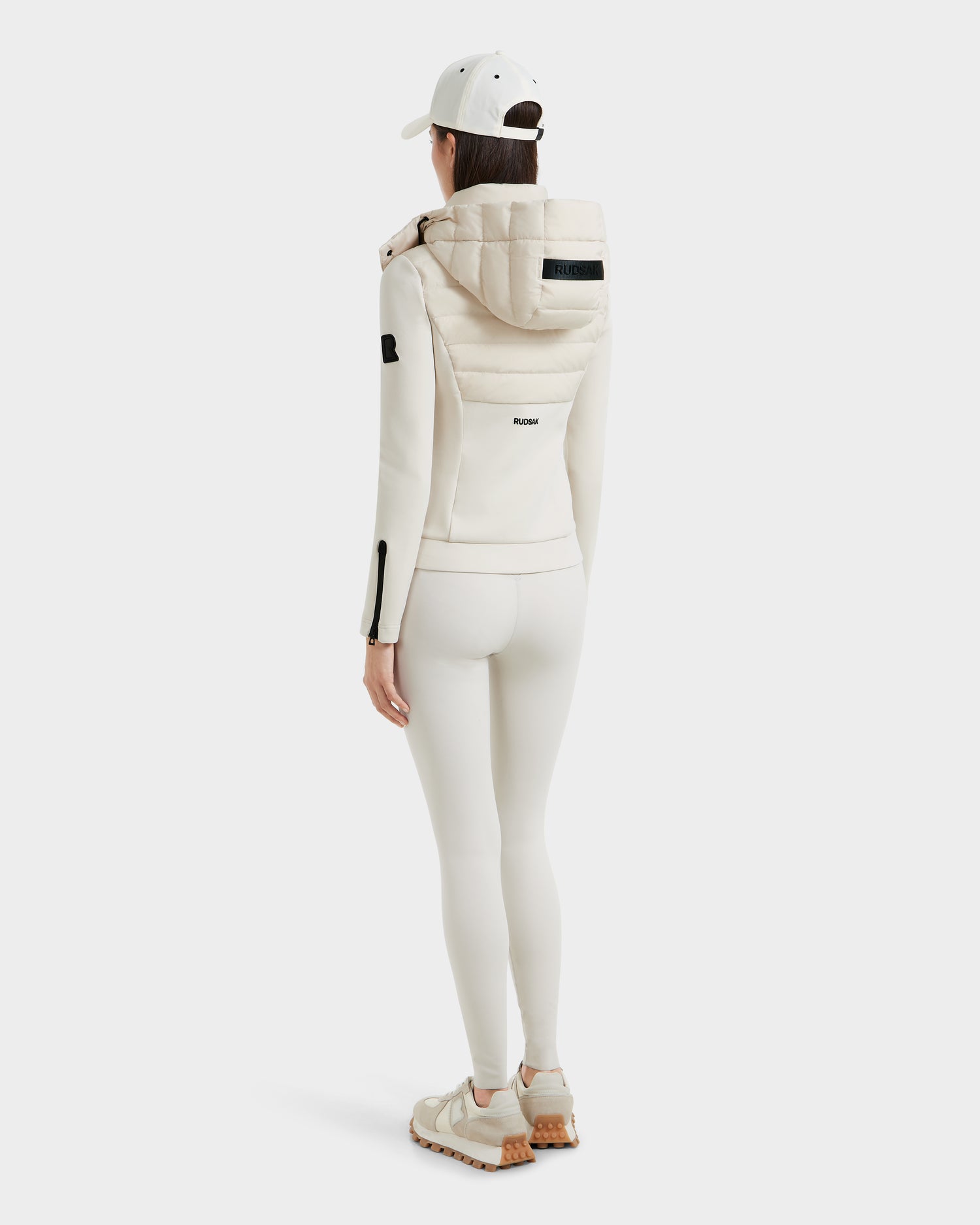 A woman dressed in all-white Rudsak MABS - 8125055 Vanilla activewear, including a lightweight jacket and fitted leggings, sneakers, and a baseball cap, stands facing away against a plain white background.