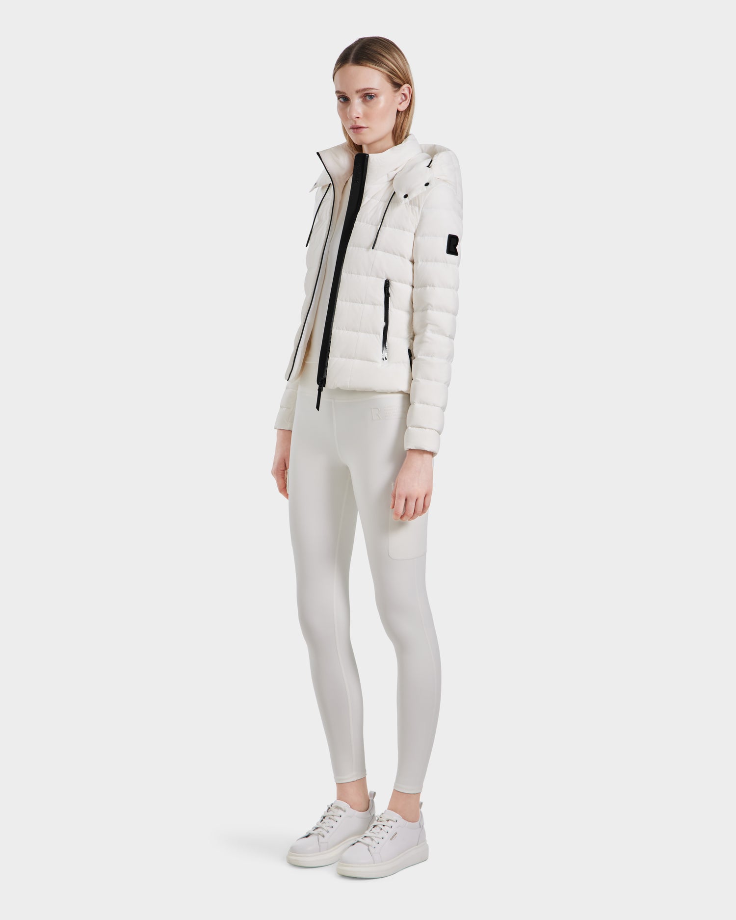 A woman stands against a plain light background, wearing the Rudsak MELISSA - 8124031 Cotton White lightweight down puffer, paired with white fitted pants and sneakers. She has straight, shoulder-length hair and a neutral expression.