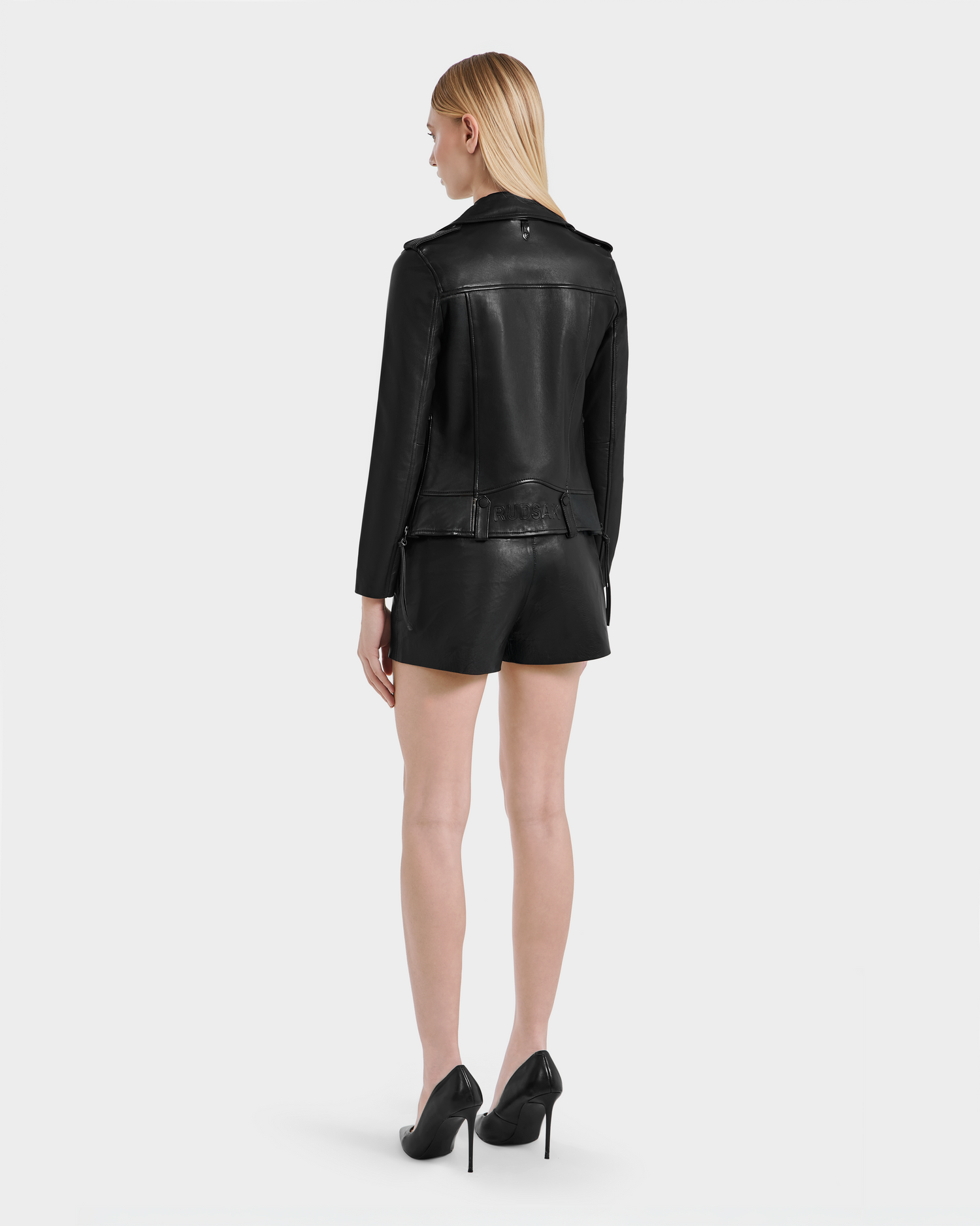 A woman with straight blonde hair stands facing away, wearing the Rudsak MERGO - 8122009 BLACK lambskin leather jacket, paired with black leather shorts and high heels against a plain white background.