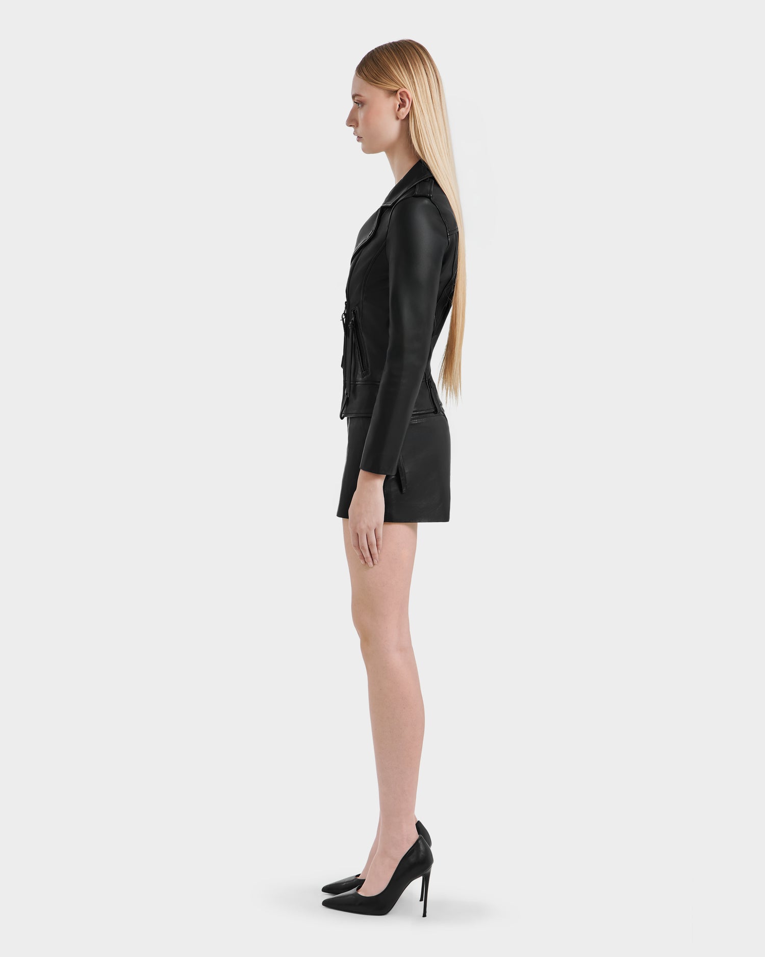 A woman with long straight blonde hair stands in profile against a plain white background, wearing the Rudsak MERGO - 8122009 BLACK lambskin leather biker jacket, a short black skirt, and black high-heeled shoes.