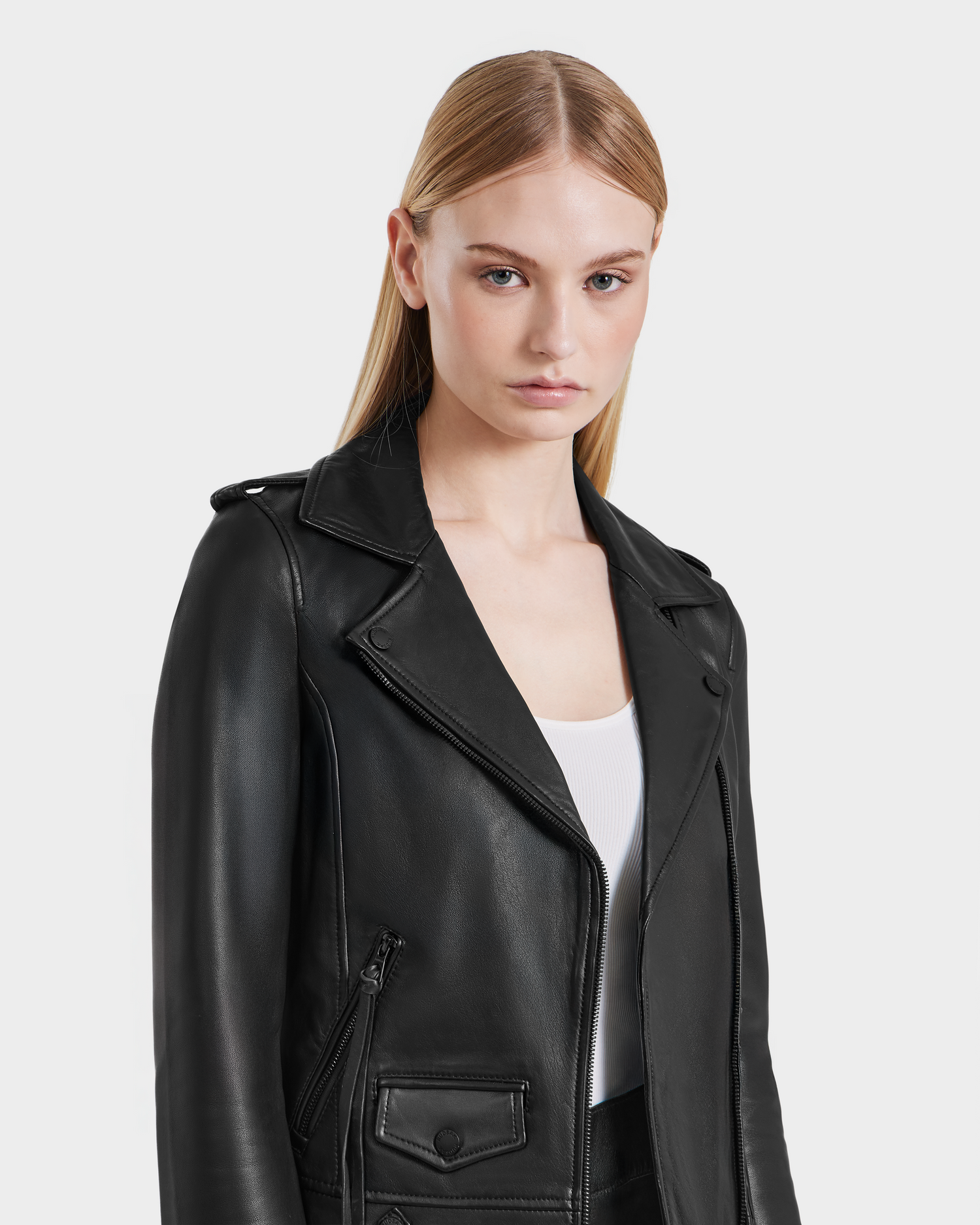 A woman with straight blonde hair wears the Rudsak MERGO - 8122009 BLACK lambskin leather biker jacket over a white top, standing against a plain light background and looking directly at the camera with a neutral expression.
