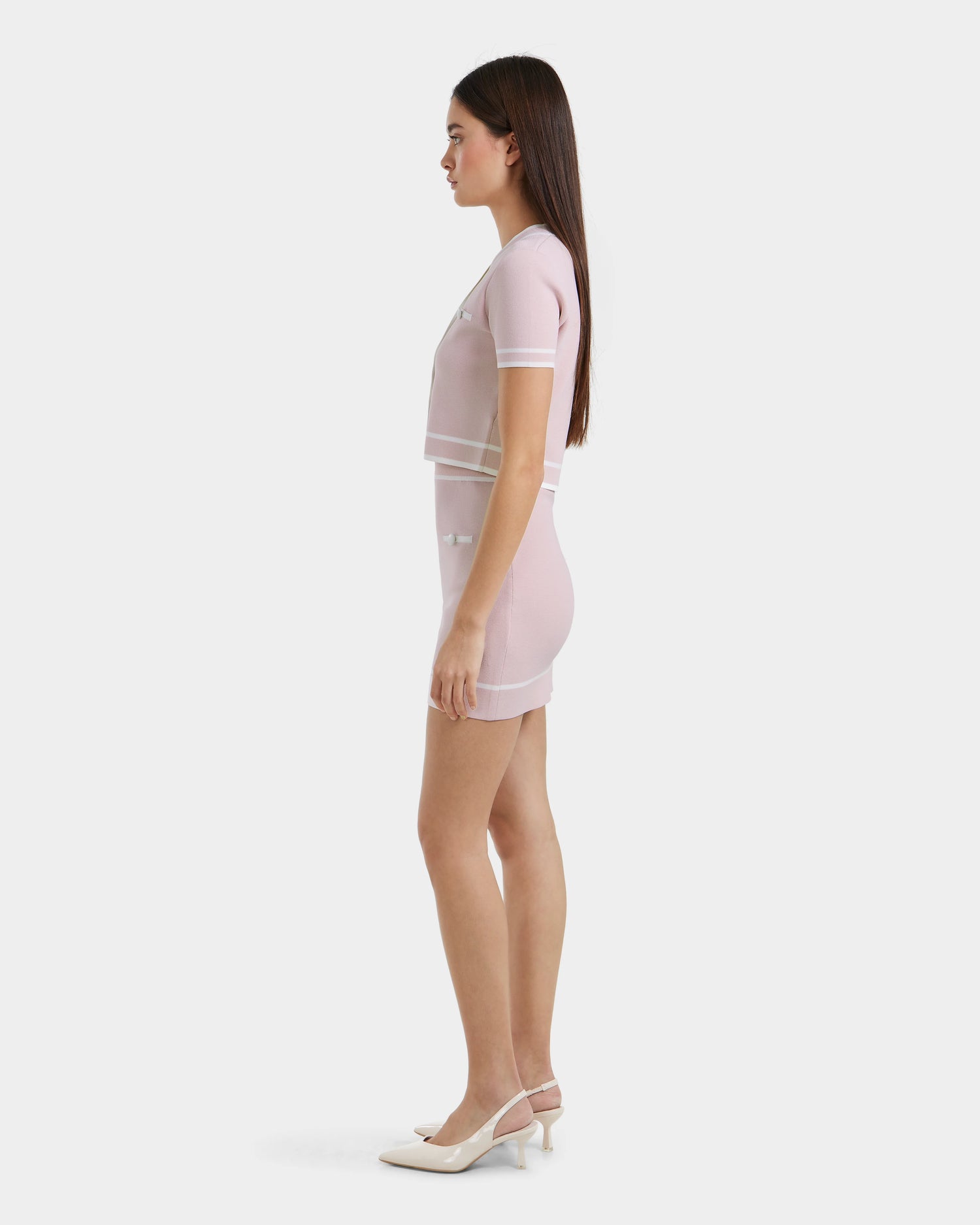 A woman stands in profile against a plain white background, wearing the Rudsak NAPA - 8525083 Light Pink fitted knit mini skirt set with white trim and short sleeves, paired with white high-heeled shoes.
