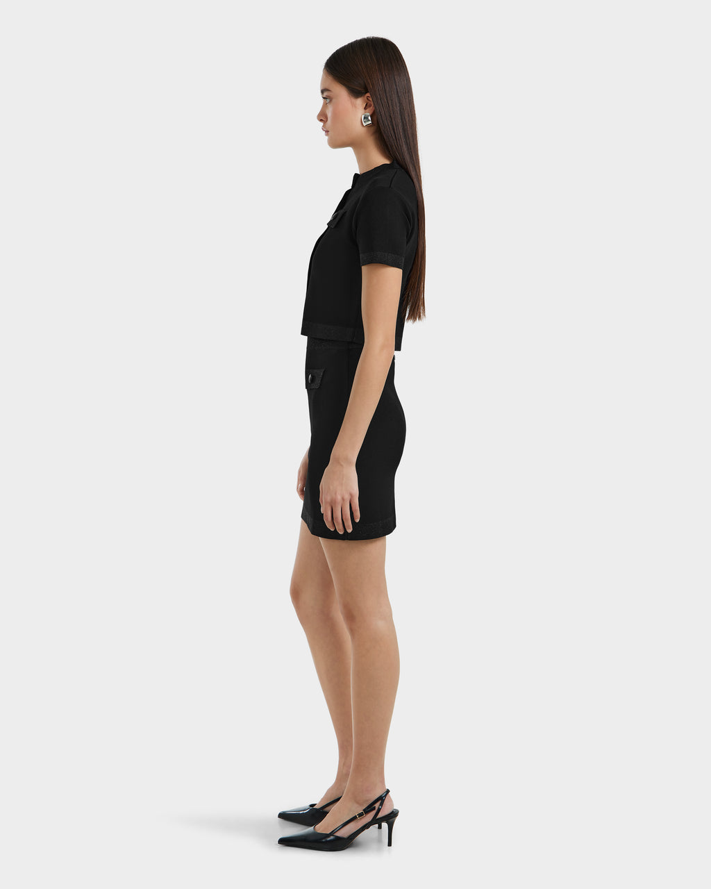A woman in profile stands against a light gray background, wearing the Rudsak PANAMA - 8525075 Black womens fitted knit mini skirt with chic button accents, a black top, slingback heels, statement earrings, and straight hair down.