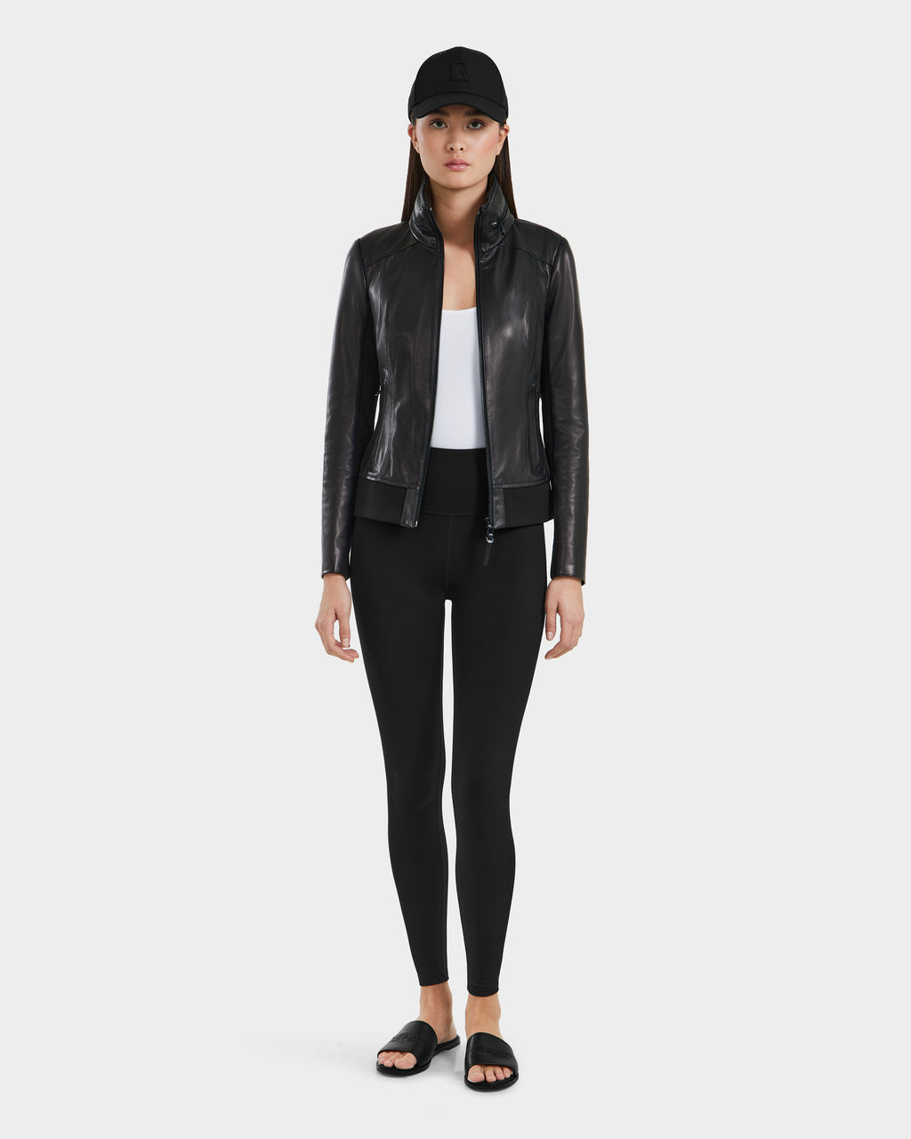 A woman faces forward against a white background, wearing the Rudsak REBECCA - 8125066 Black moto-inspired bomber lamb leather jacket with a lightweight hideaway hood, paired with black leggings, a white top, cap, and slide sandals.