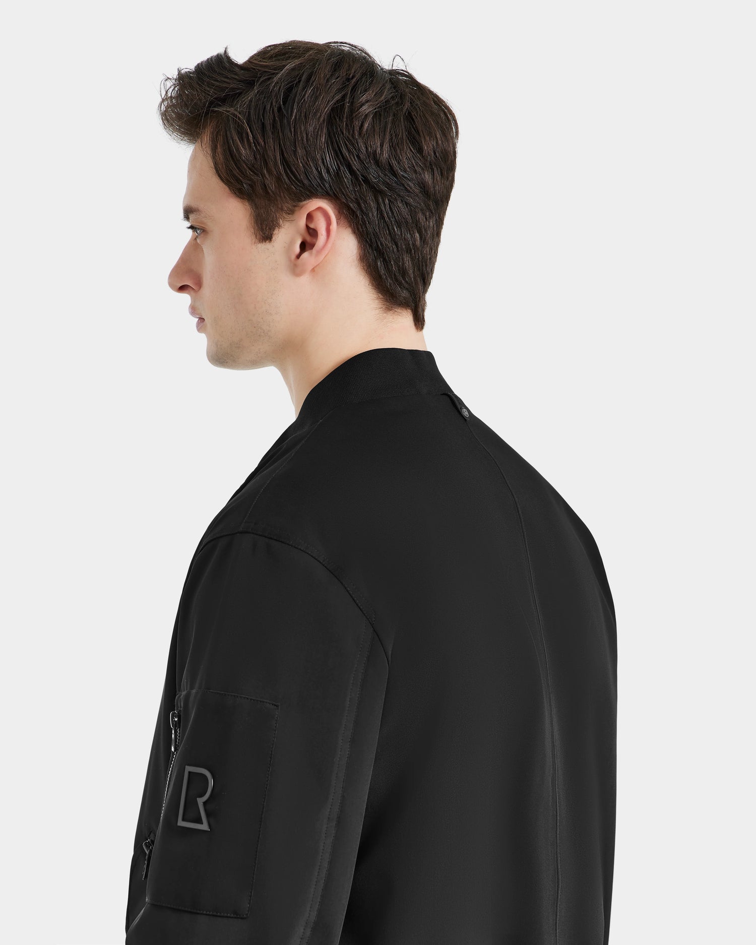 A young man with short brown hair is seen from the side and back, wearing the Rudsak RICO - 6124021 Black bomber jacket featuring a patch on the sleeve, set against a plain light background.