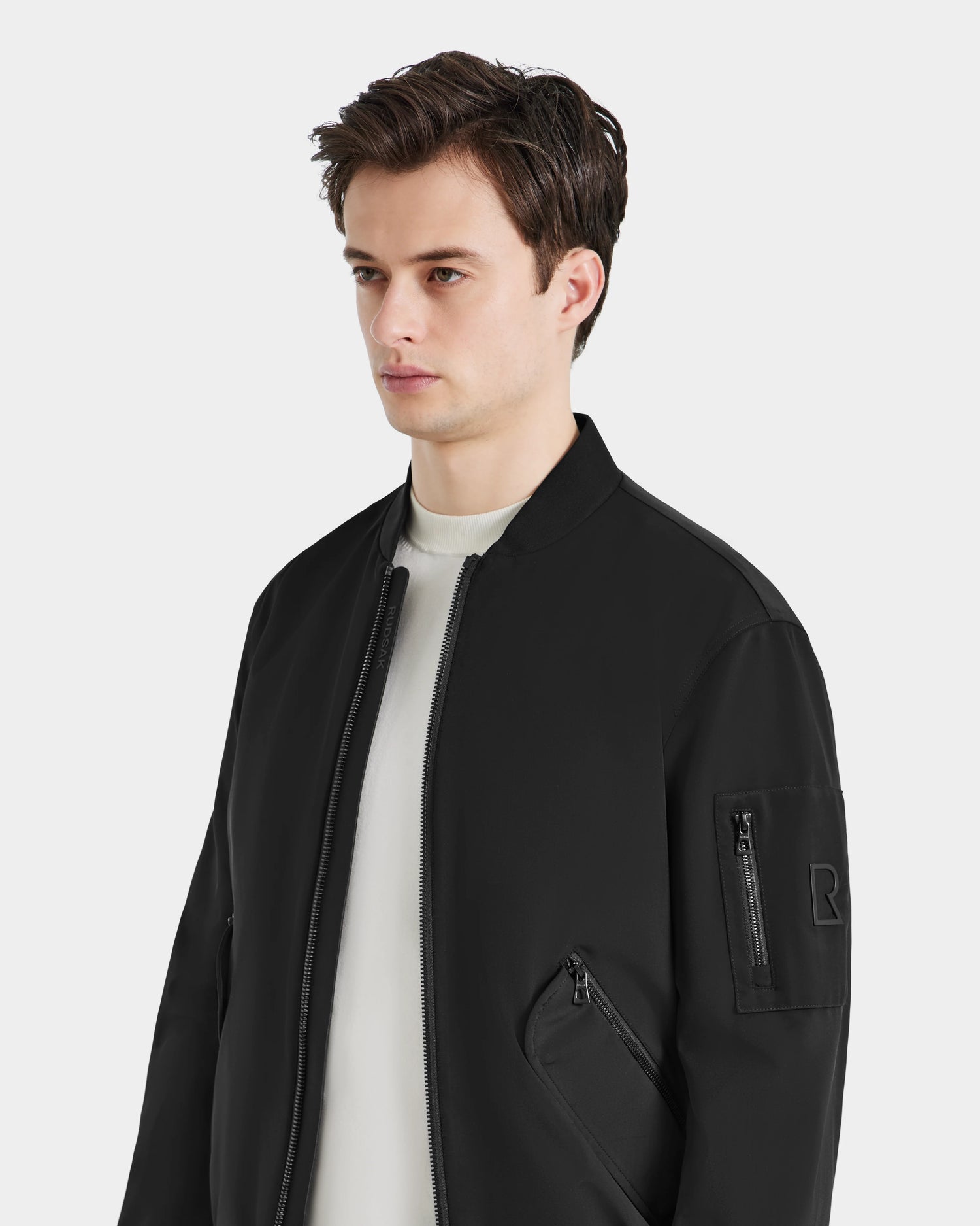 A young man with short brown hair models the RICO - 6124021 Black mens bomber jacket by Rudsak over a white shirt, standing against a plain light background and gazing slightly to the side.
