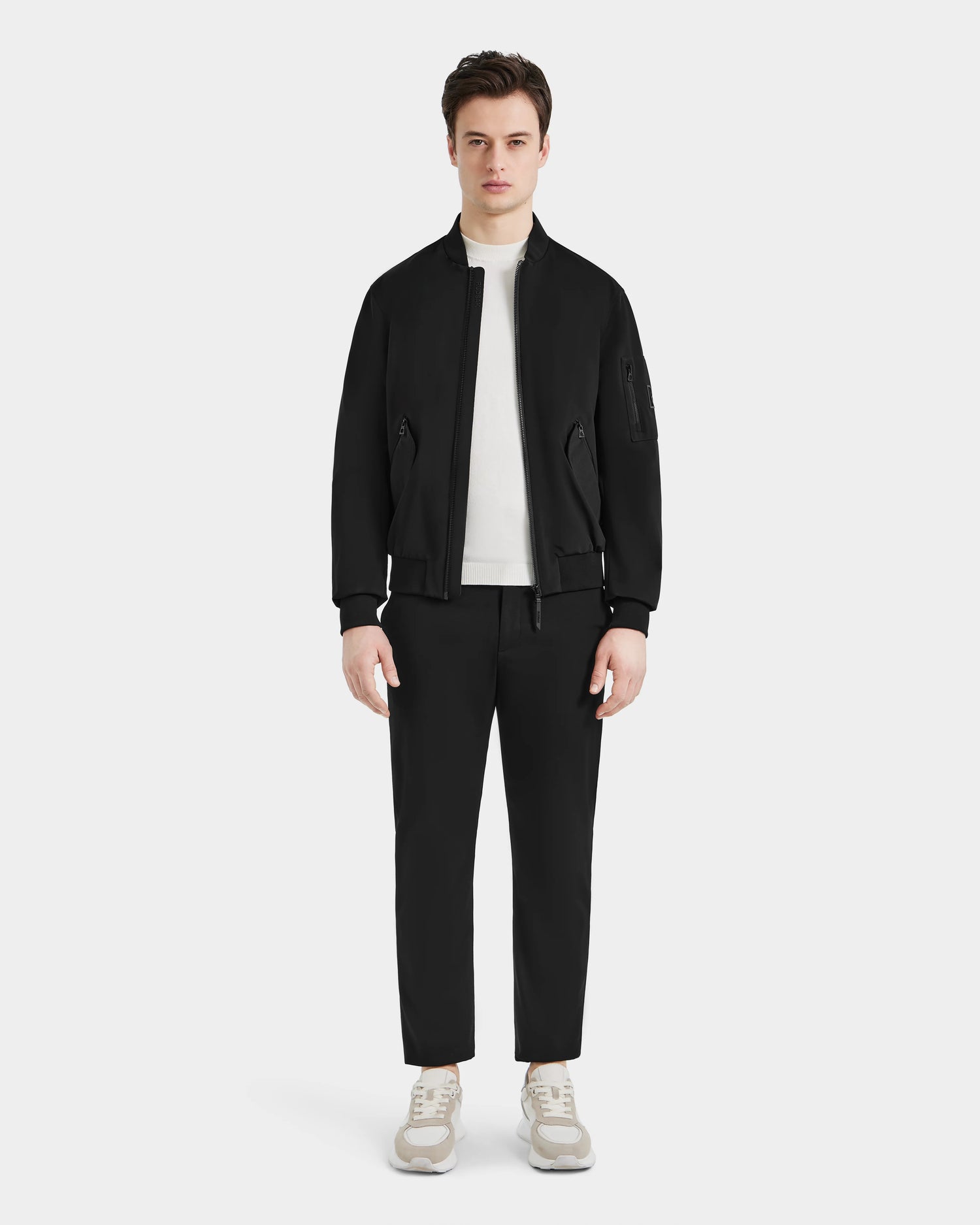 A man faces forward wearing the Rudsak RICO - 6124021 Black classic bomber jacket, styled with black pants, a white turtleneck sweater, and white sneakers against a plain light background.