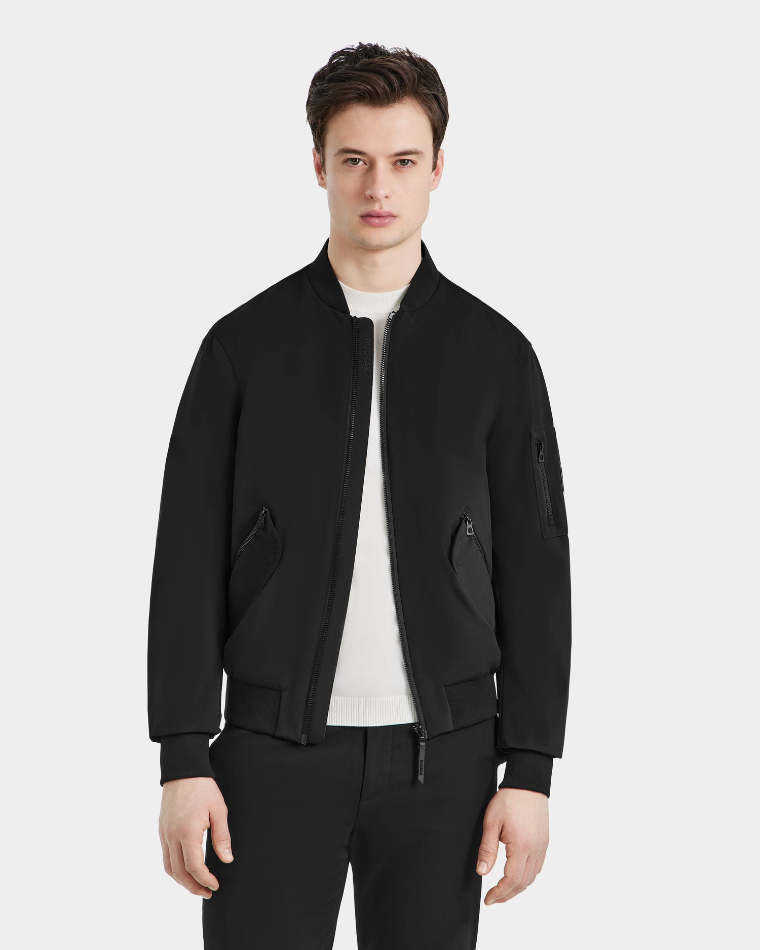A young man faces forward, embodying urban sophistication in the Rudsak RICO - 6124021 Black mens bomber jacket, styled over a white shirt and black pants against a plain light gray background.