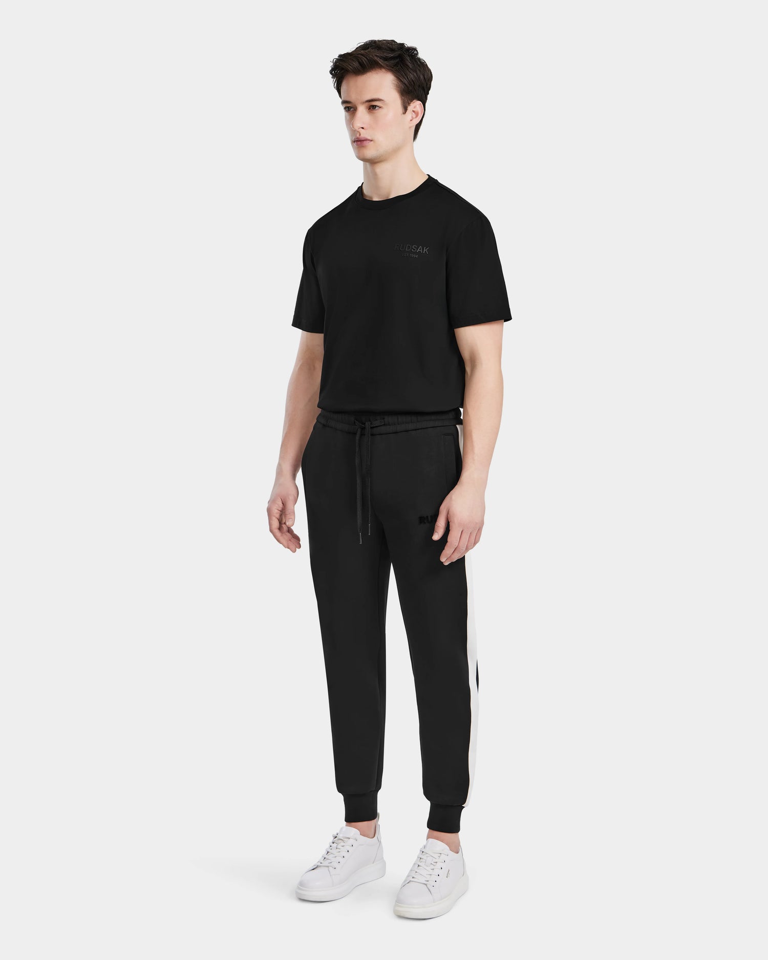 A man stands facing forward, wearing a black t-shirt, Rudsak RYLAN - 6525009 Black men’s joggers with white side stripes and white sneakers, against a plain white background.