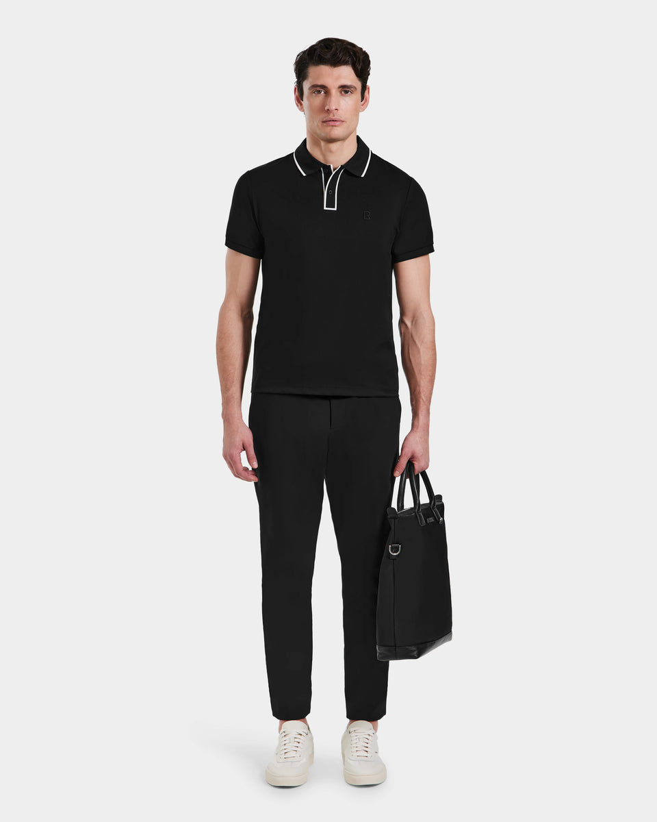 A man stands facing forward against a plain white background, wearing the Rudsak XAVIER - 6524027 Black mens polo shirt with white trim, paired with black pants and white sneakers, holding a black tote bag in his left hand.