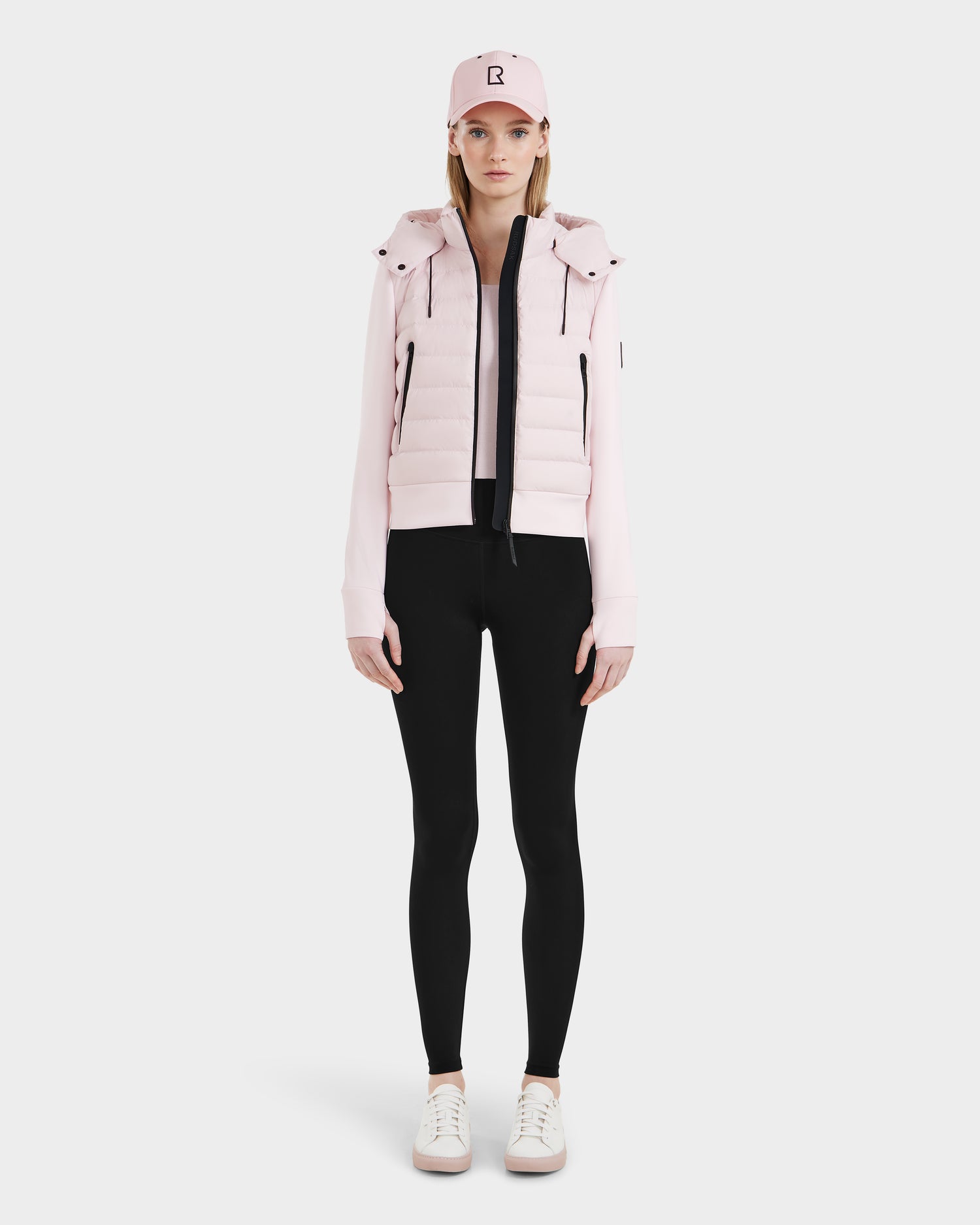 A woman faces forward against a white background, wearing the Rudsak CASSA - 8125057 light pink cropped puffer jacket, styled with black leggings, white sneakers, and a pink baseball cap—ideal for transitional weather.