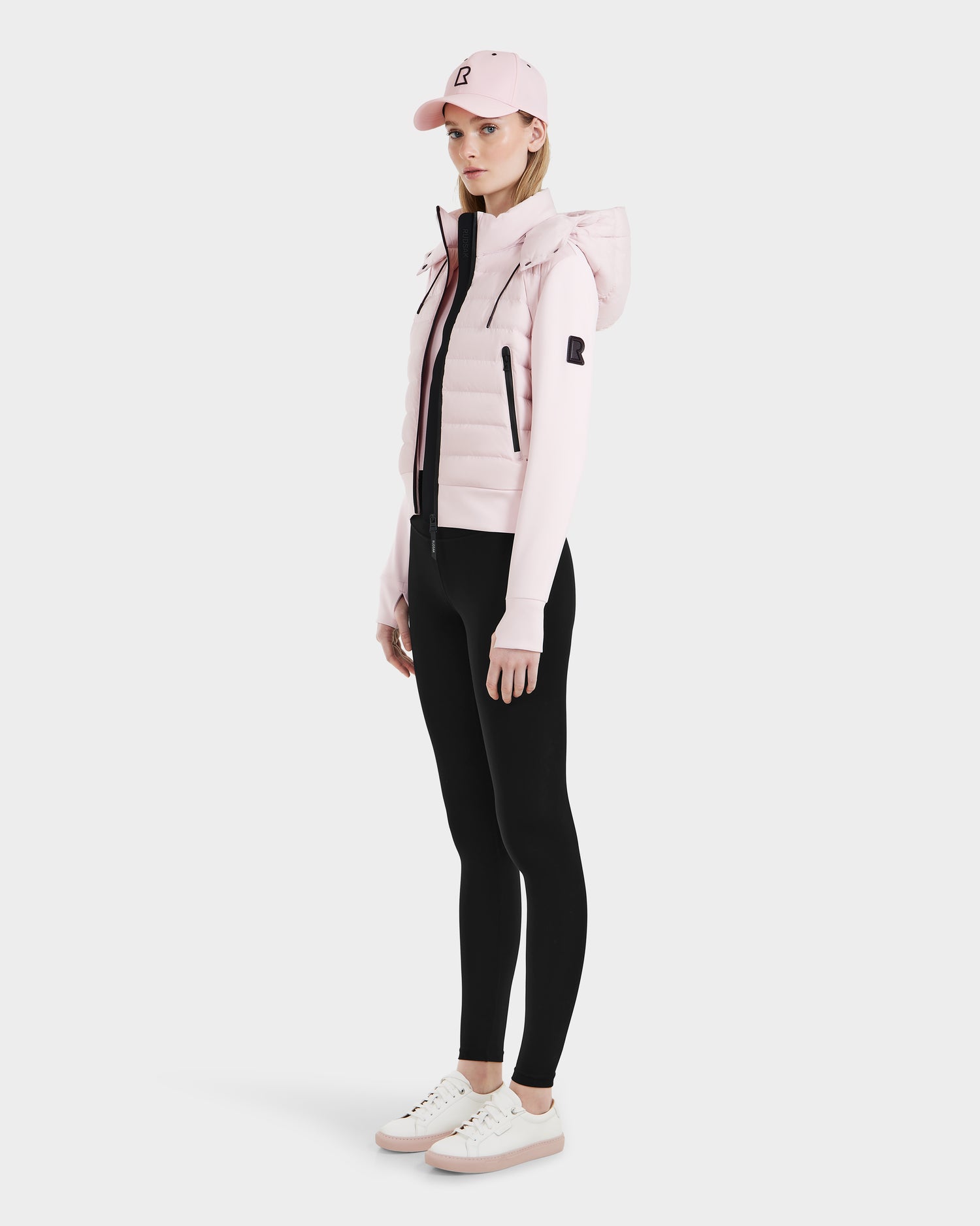 A woman stands against a plain background wearing the Rudsak CASSA - 8125057 lightweight cropped jacket in light pink, paired with black leggings and white sneakers. She faces slightly to the side with her arms relaxed at her sides.