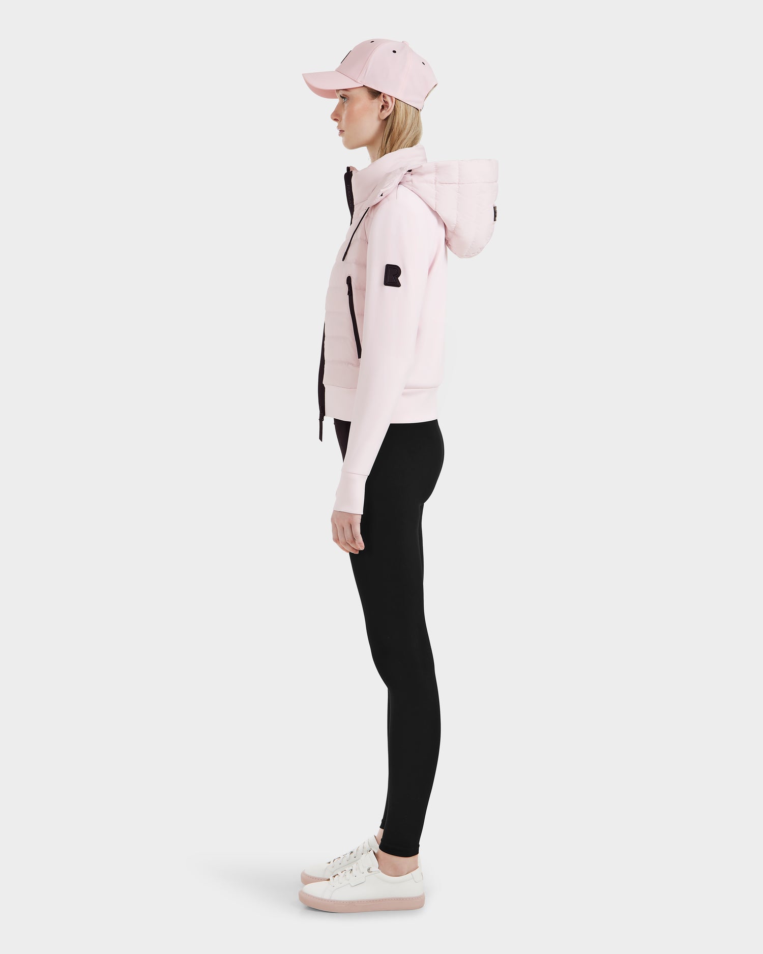 A woman in profile poses against a white background, wearing the Rudsak CASSA – 8125057 Light Pink jacket over a hoodie, with a matching pink cap, black leggings, and white sneakers with pink soles.