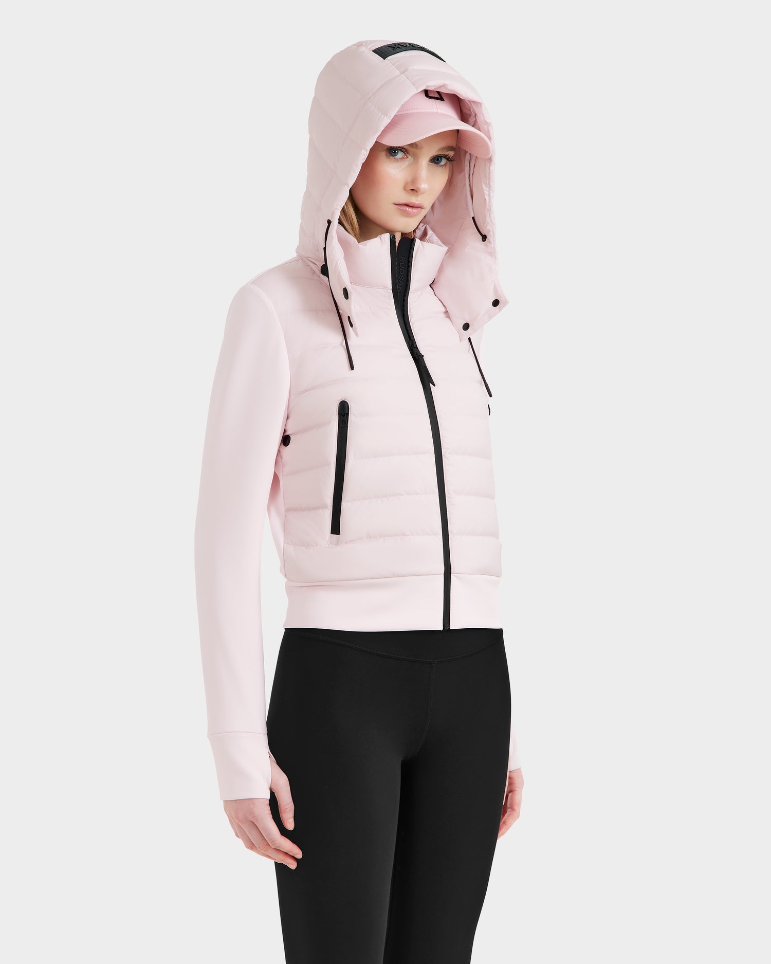 A woman wears the Rudsak CASSA - 8125057 lightweight cropped jacket in light pink, paired with a pink cap and black leggings, standing against a plain white background and gazing slightly to the side.