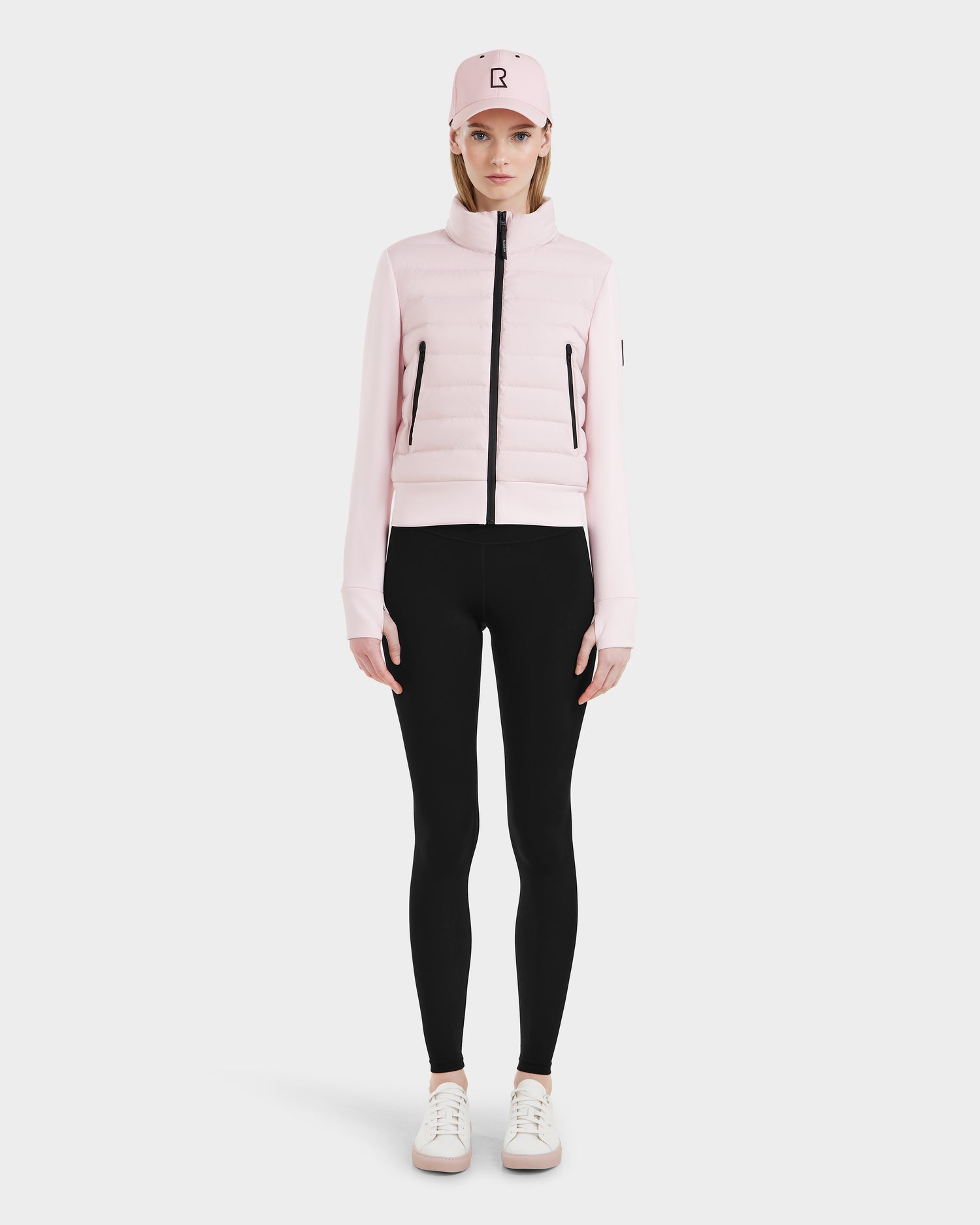 A woman faces forward in a light pink Rudsak CASSA - 8125057 cropped jacket, black leggings, white sneakers, and a matching pink baseball cap against a plain white background.