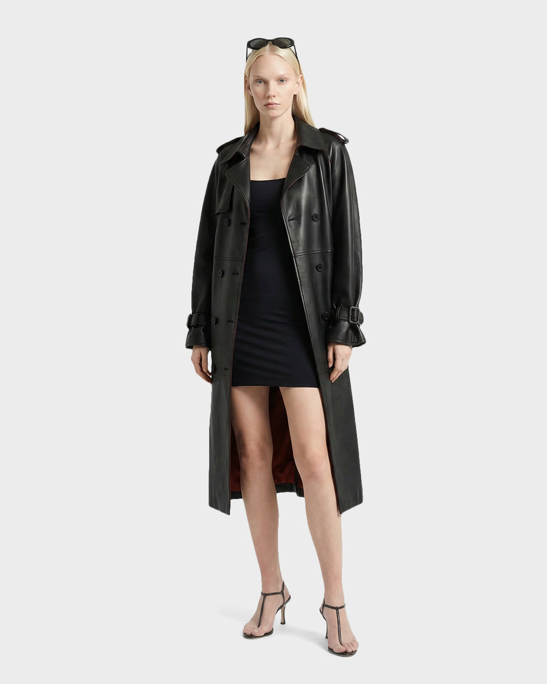 A woman stands facing forward against a plain light background, wearing the Rudsak CASSIE L-B - 8125553 Black trench coat over a short black dress and high-heeled sandals, with sunglasses on her head.