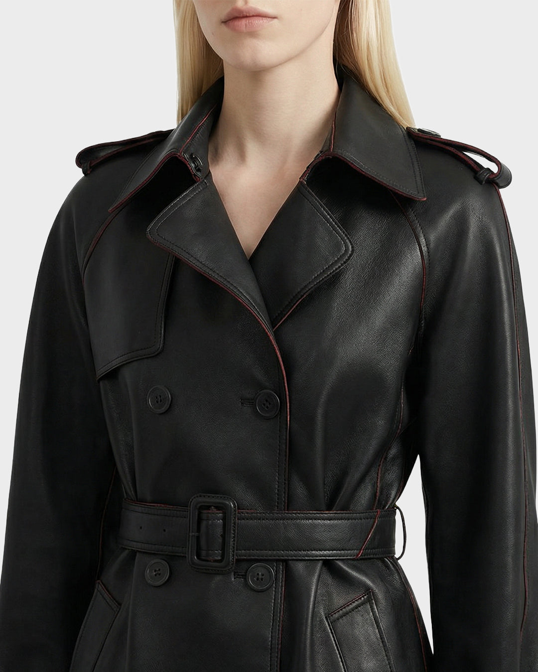 A woman wears the Rudsak CASSIE L-B - 8125553 Black women's leather trench coat, featuring a belted waist, buttoned front, epaulets, and subtle red seam piping. Only her lower face and upper body are visible.