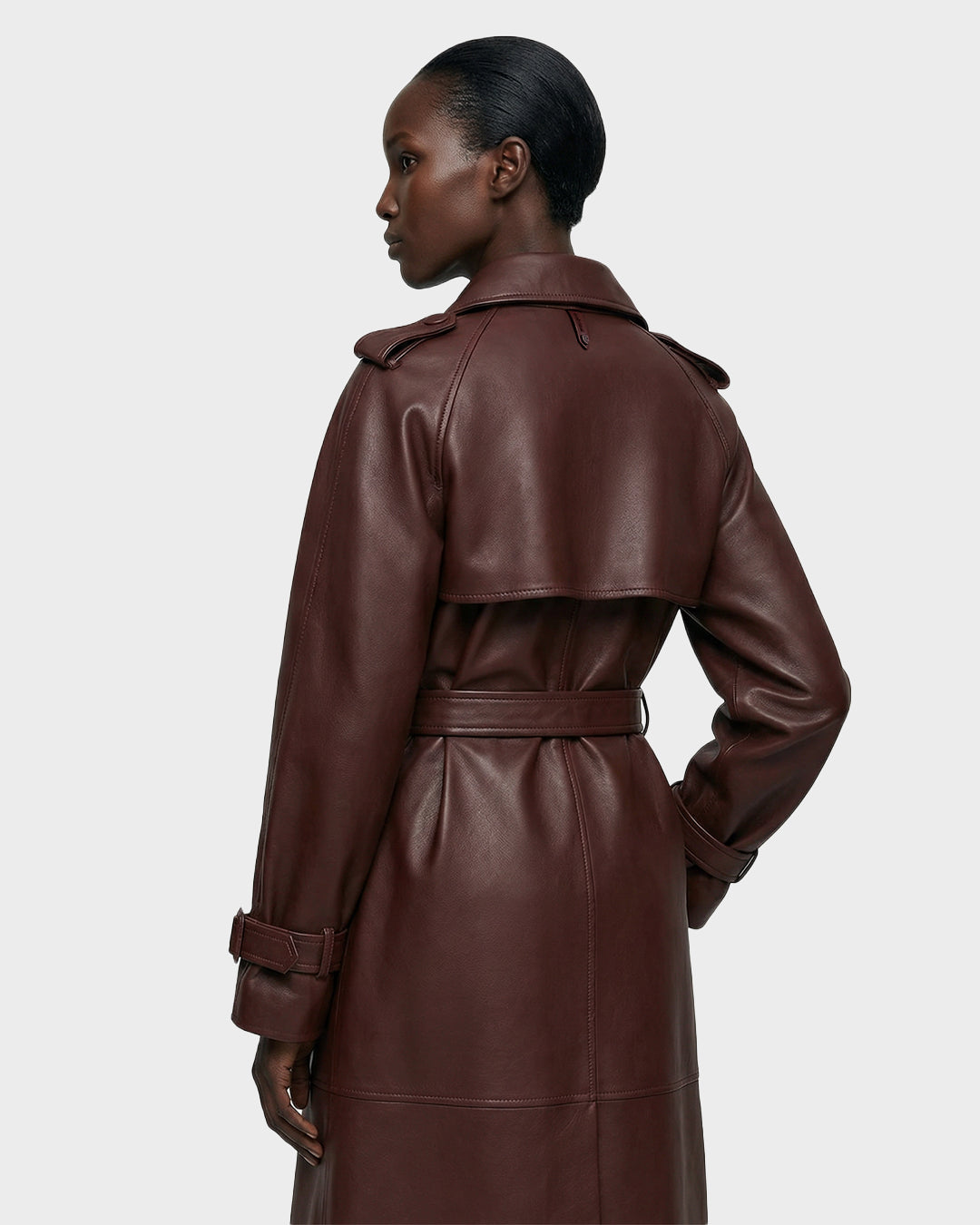 A woman with dark skin and a low bun is seen from behind, wearing the Rudsak CASSIE L-B - 8125553 Rust women’s bonded leather trench coat with belt, double-breasted design, epaulets, and storm flap against a plain light grey background.