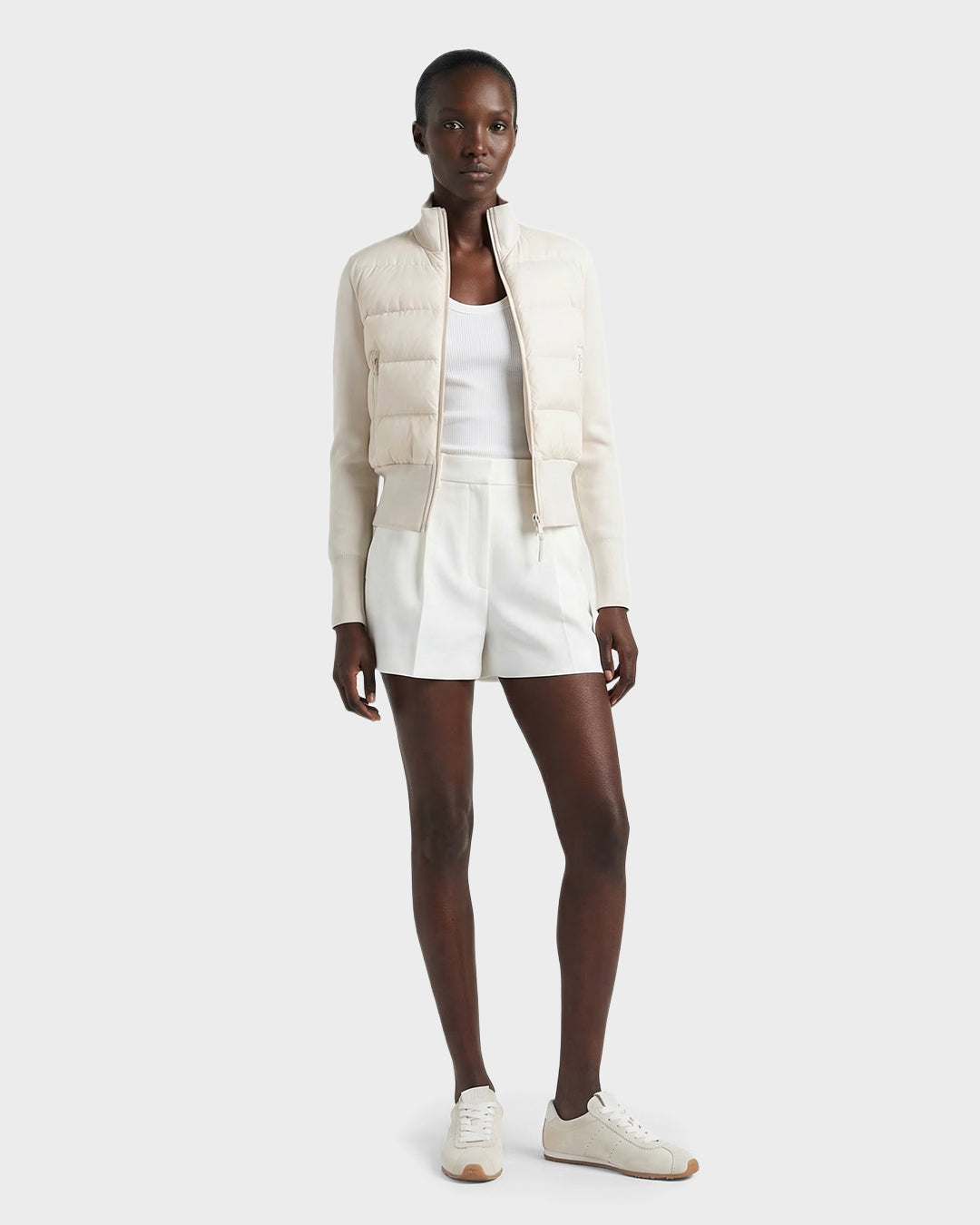 A woman wears the Rudsak CLAREMONT - 8125502 Vanilla hybrid down jacket, styled over a white tank top, high-waisted shorts, and white sneakers against a plain light backdrop.