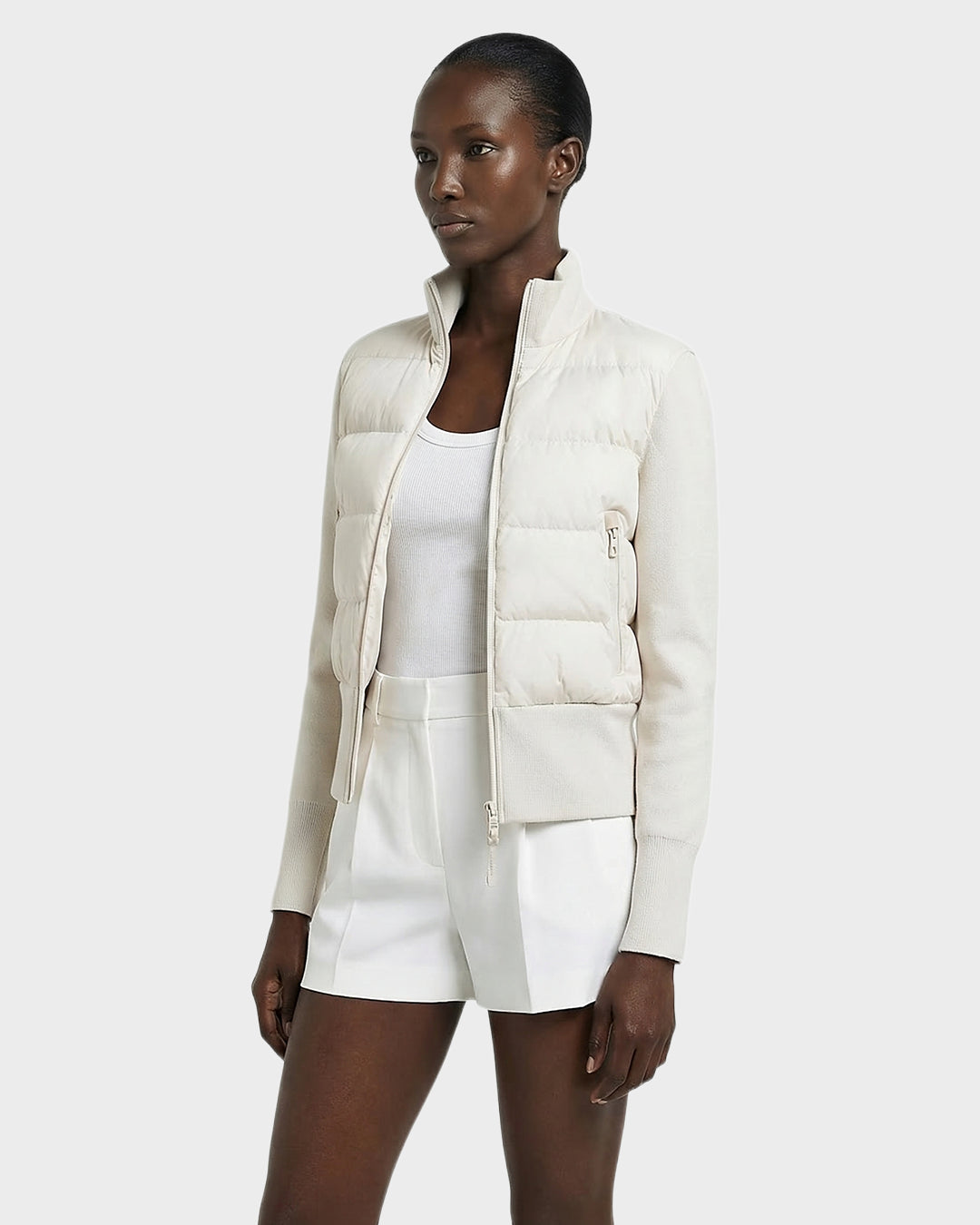 A woman models the Rudsak CLAREMONT - 8125502 Vanilla women’s hybrid down jacket with RDS-certified down over a white tank top and shorts, standing against a plain light background and highlighting its lightweight warmth.