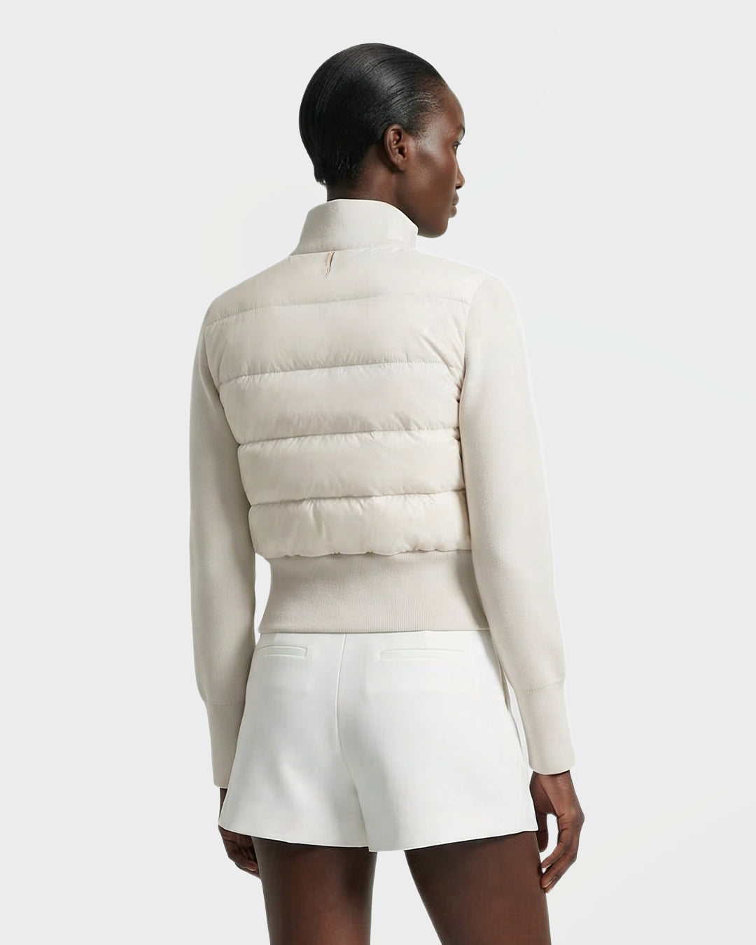 A woman stands with her back to the camera, wearing Rudsak’s CLAREMONT - 8125502 Vanilla hybrid down jacket and white shorts, her slicked-back hair and the light background emphasizing the jacket’s lightweight warmth.