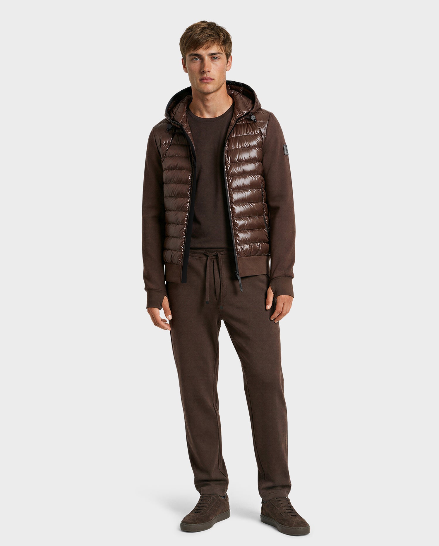 A man faces forward wearing the Rudsak DEE S - 6125684 Espresso men’s lightweight down jacket with hood over a matching brown sweatshirt and drawstring pants, paired with brown sneakers—an ideal urban layer against a plain light backdrop.