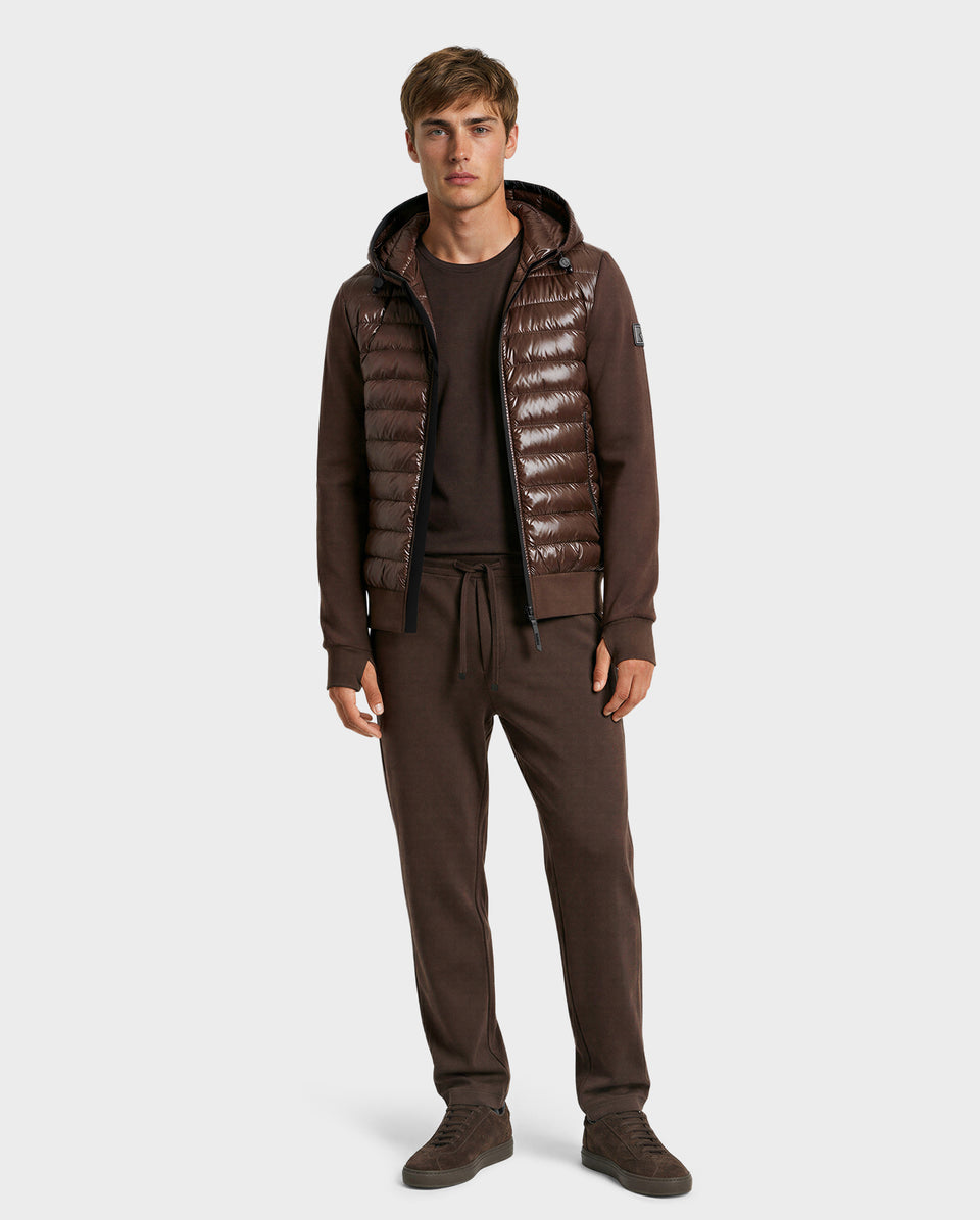 A man faces forward wearing the Rudsak DEE S - 6125684 Espresso men’s lightweight down jacket with hood over a matching brown sweatshirt and drawstring pants, paired with brown sneakers—an ideal urban layer against a plain light backdrop.