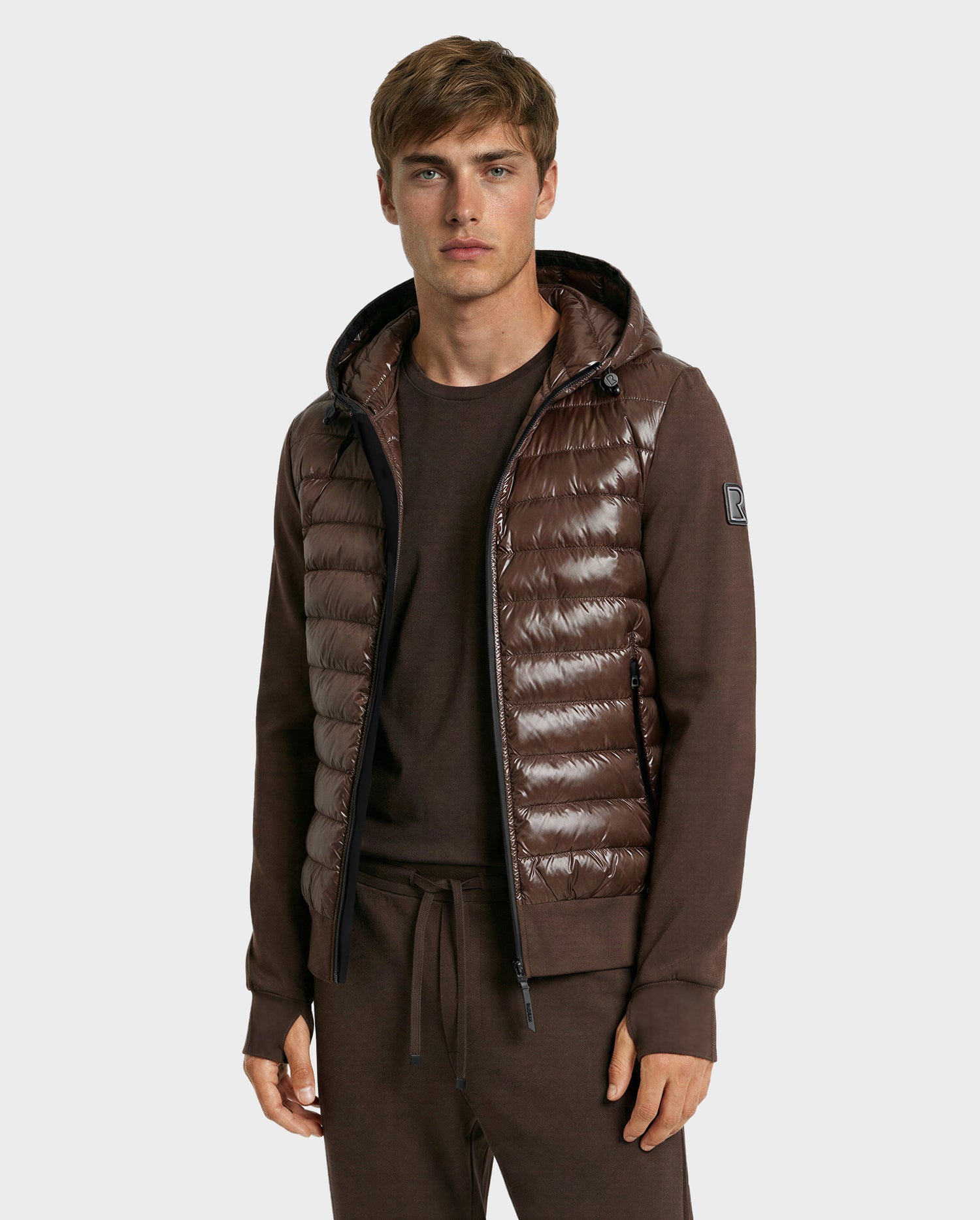 A young man faces forward against a plain light background, wearing the Rudsak DEE S - 6125684 Espresso men's lightweight down jacket over a brown shirt and pants. The jacket is shiny, quilted, and filled with RDS-certified duck down.