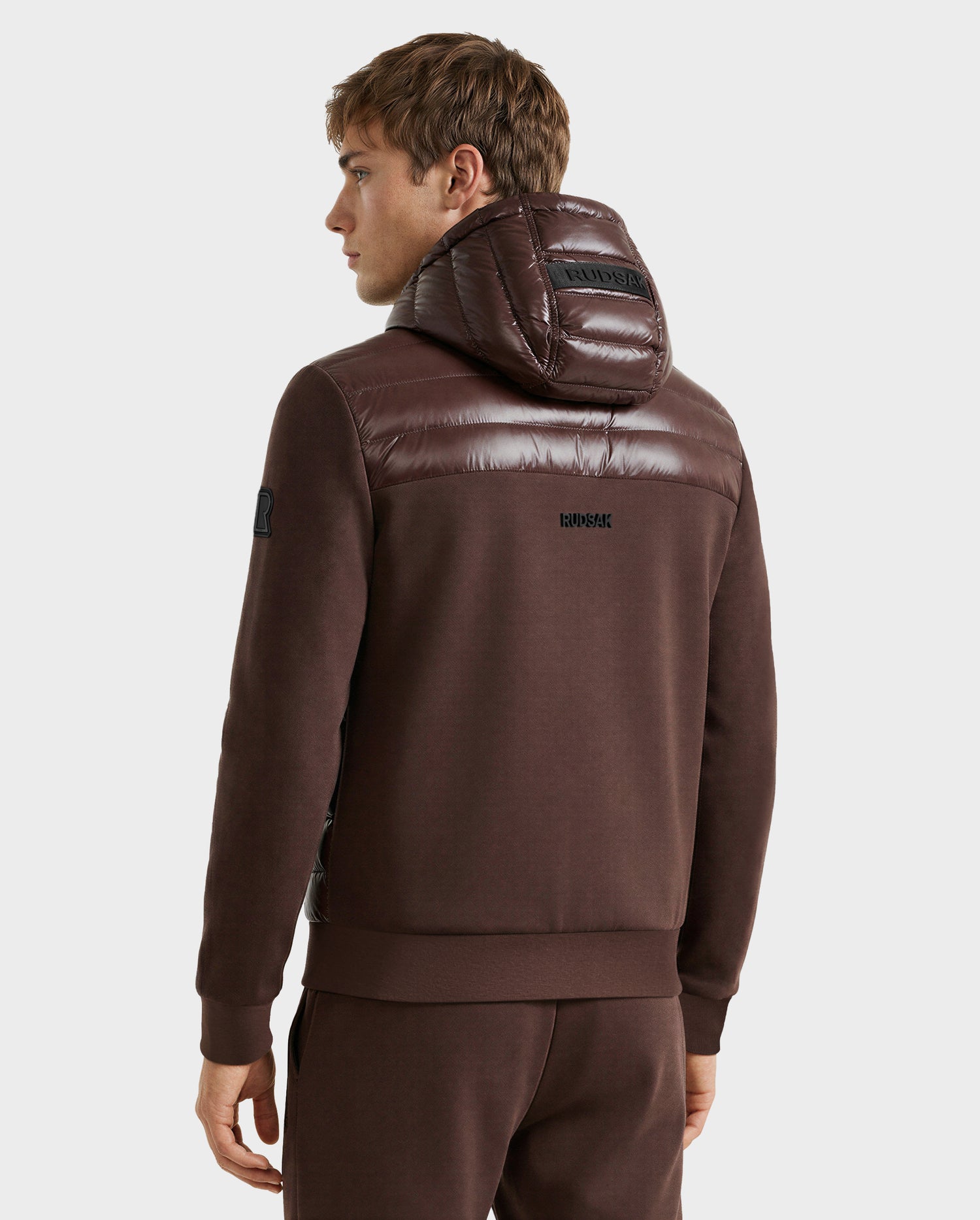 A man is seen from behind wearing the Rudsak DEE S - 6125684 Espresso lightweight down jacket with shiny, quilted fabric on the hood and shoulders—perfect for city style. He pairs it with matching brown pants against a plain white background.