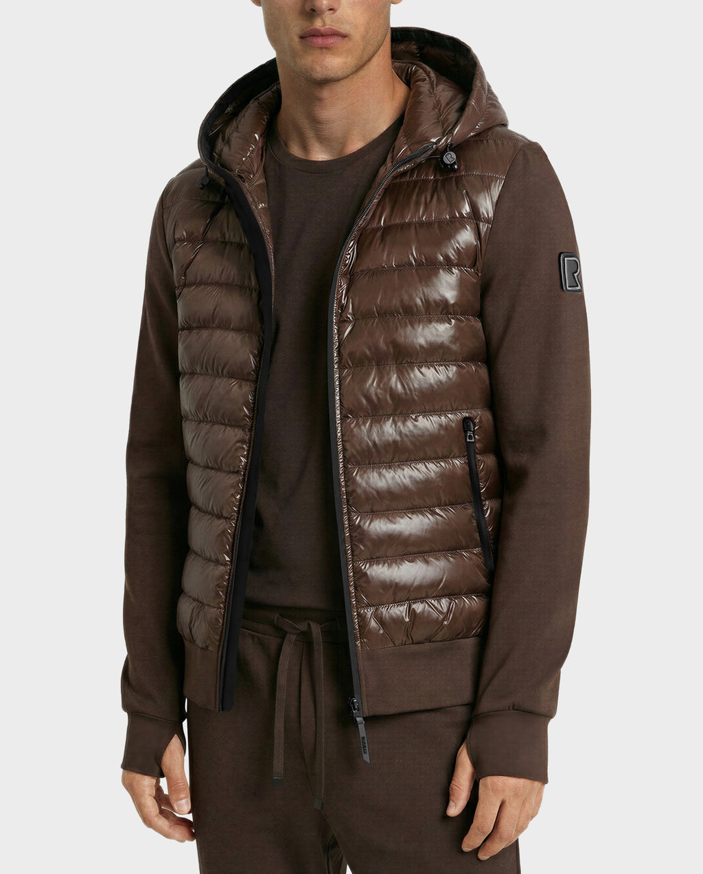 A man wears the Rudsak DEE S - 6125684 Espresso shiny brown urban puffer vest with a hood, filled with RDS-certified duck down and zippered pockets, over a matching long-sleeve shirt featuring an upper sleeve patch and drawstring pants.