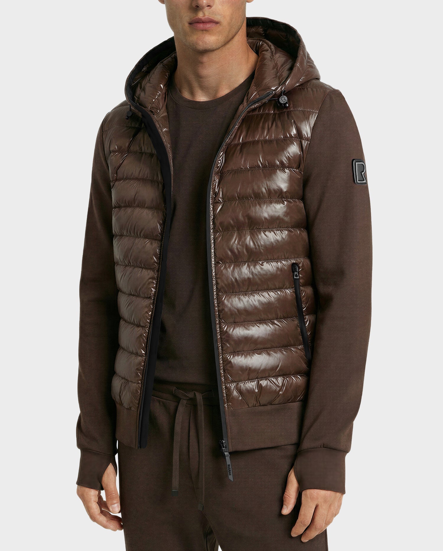 A man wears the Rudsak DEE S - 6125684 Espresso shiny brown urban puffer vest with a hood, filled with RDS-certified duck down and zippered pockets, over a matching long-sleeve shirt featuring an upper sleeve patch and drawstring pants.