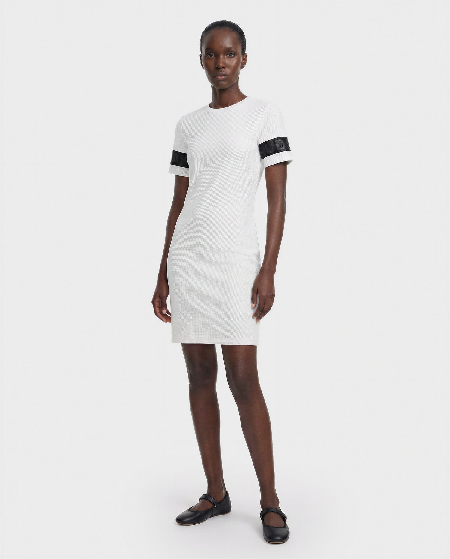 A woman stands against a plain white background wearing the Rudsak ELAINE - L523029 Optic White short-sleeved dress with black bands on the sleeves and black Mary Jane shoes.