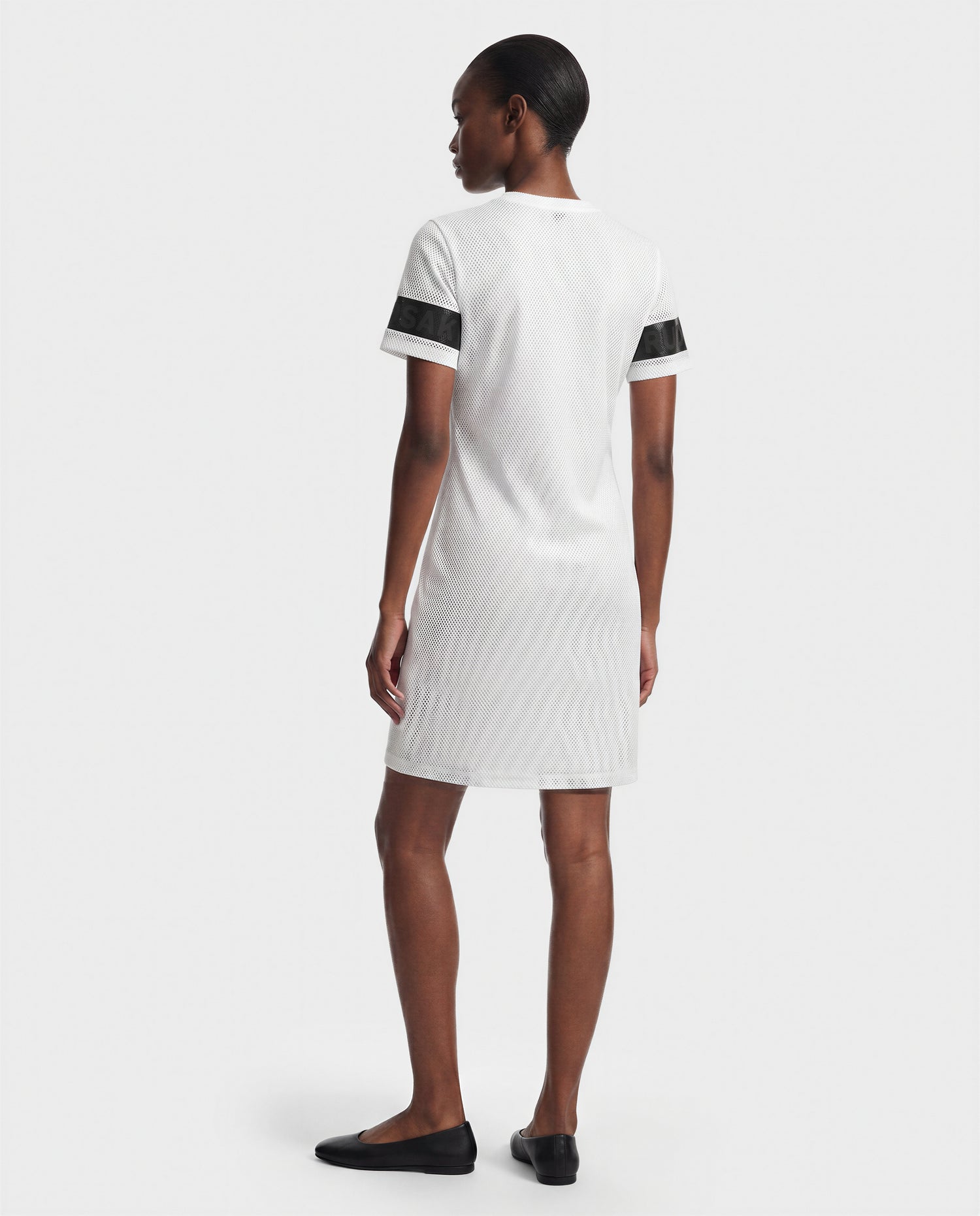 A woman faces away in the ELAINE - L523029 Optic White short-sleeve dress by Rudsak, featuring black sleeve stripes and paired with black flats, set against a plain light background.