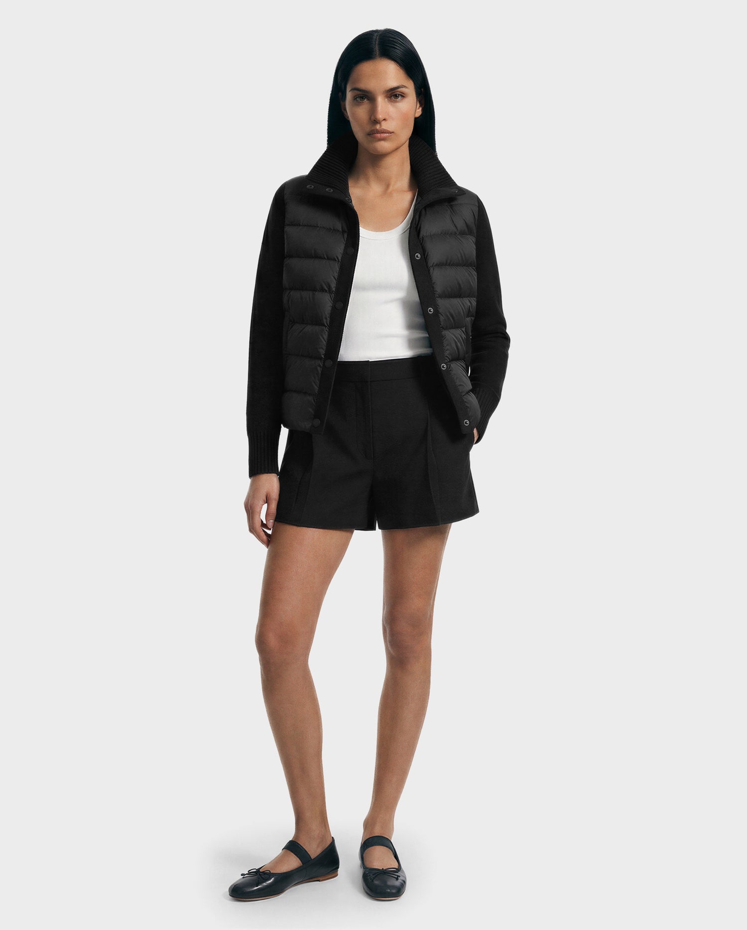 A woman faces forward in the Rudsak ELLIE - 8125635 Black quilted down jacket, layered over a white tank top, black shorts, and black flats—ideal for transitioning seasons against a plain white background.