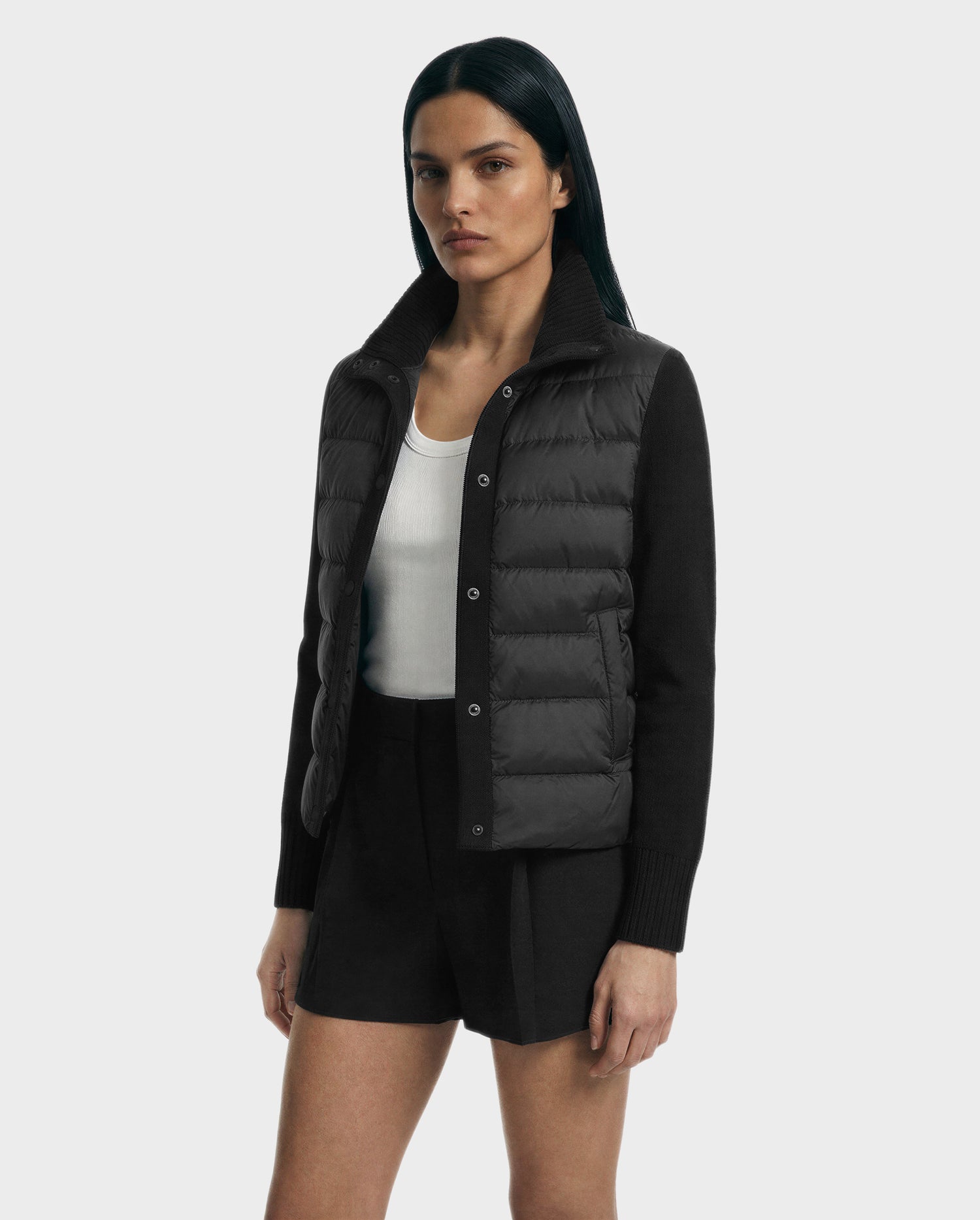 A woman with straight dark hair models the Rudsak ELLIE - 8125635 Black quilted down jacket over a white tank top and black shorts, displaying chic transitional layering against a plain light gray background.