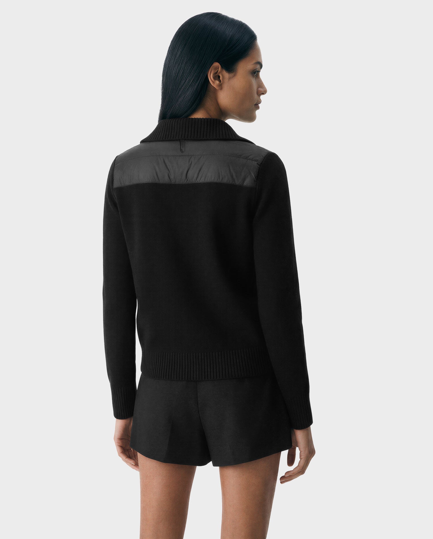 A woman with long dark hair is shown from the back wearing the ELLIE - 8125635 Black sweater and black shorts by Rudsak, featuring a shiny yoke detail—perfect for transitional layering—against a plain light gray background.