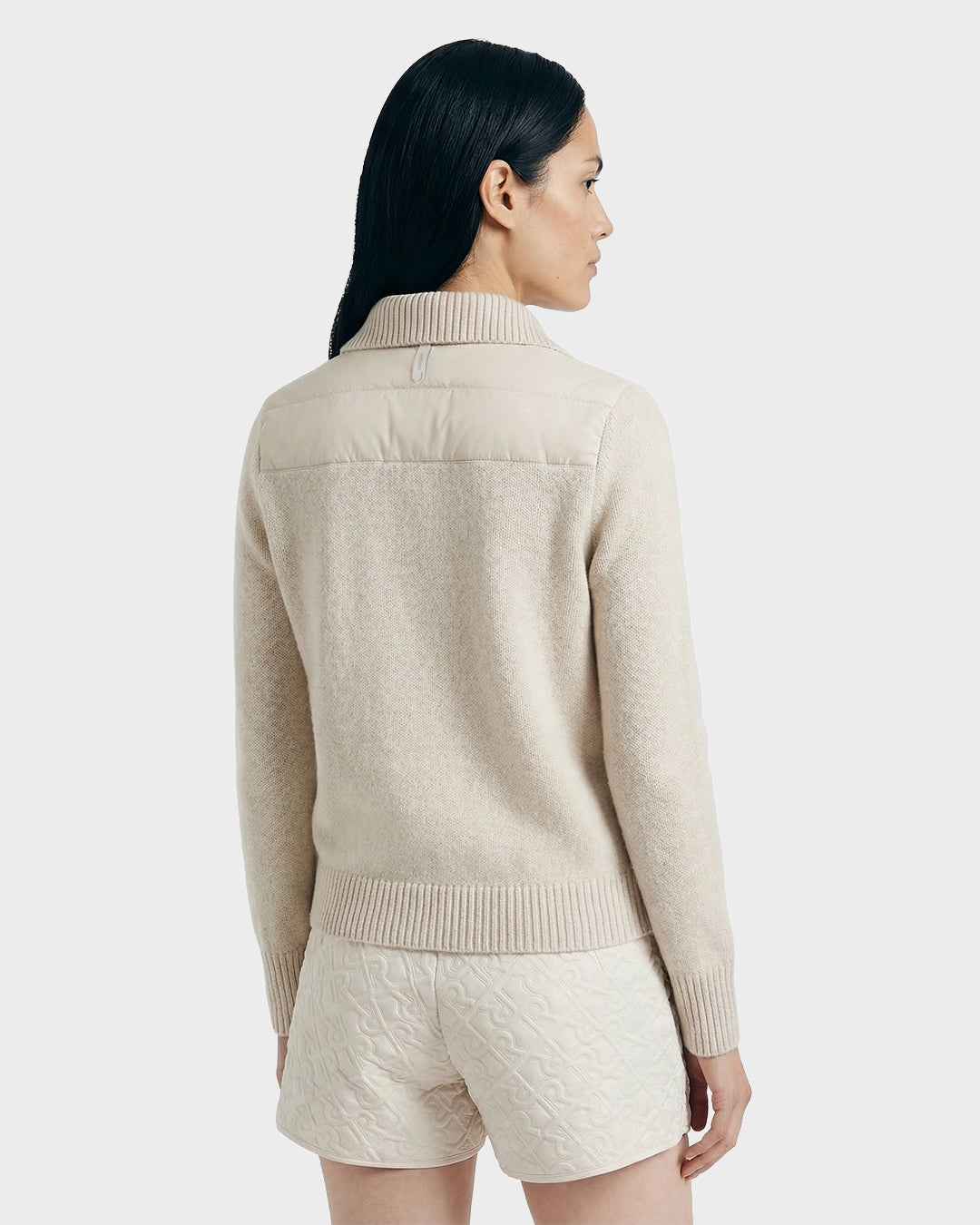 A woman with long dark hair is seen from the back, wearing Rudsak’s ELLIE - 8125635 Vanilla beige sweater with a ribbed hem and collar, paired with light quilted shorts, set against a plain light gray background.