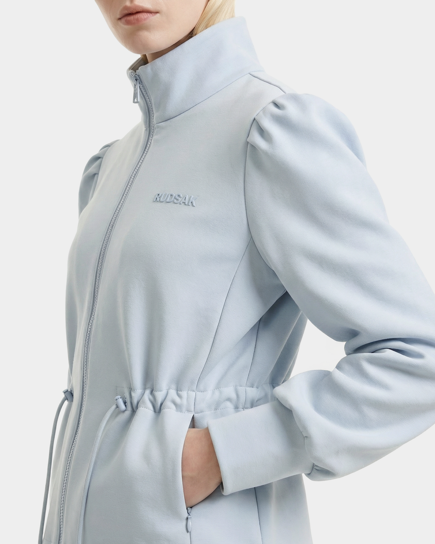 A person wears the Rudsak ELODIE - 8526039 Powder Blue zip-up sweater jacket, featuring puffed shoulders, chest embroidery, and a hand in the pocket. Made from a stretch cotton blend, it stands out against a plain white background.