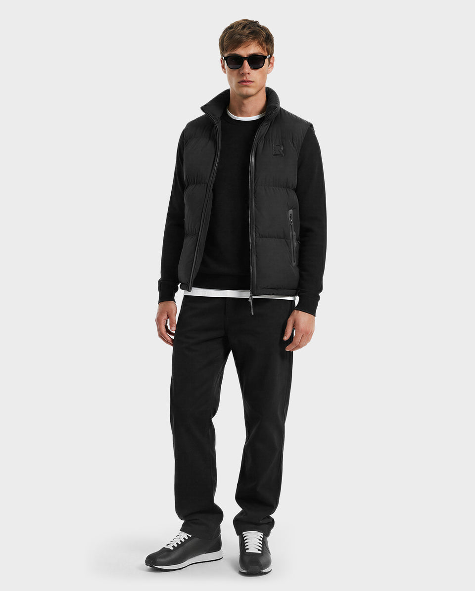 A man stands facing forward against a light gray background, showcasing effortless lightweight layering in Rudsak’s FREDDY - 6125571 Black puffer vest over a black shirt, black pants, and black sneakers with white soles.