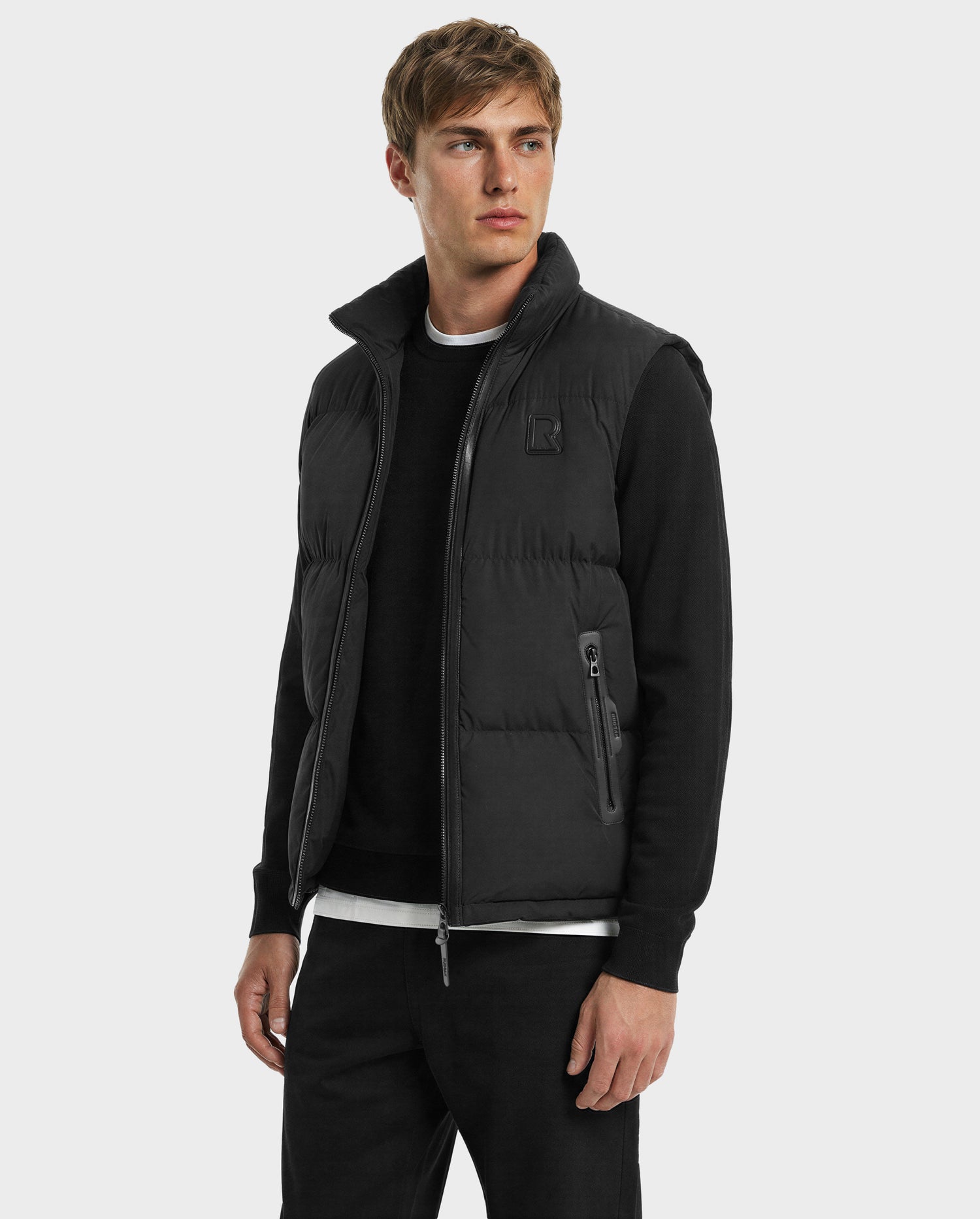 A man with short light brown hair models the Rudsak FREDDY - 6125571 Black men’s puffer vest over a black long-sleeve shirt and pants, highlighting its premium down and lightweight design against a plain light gray background.