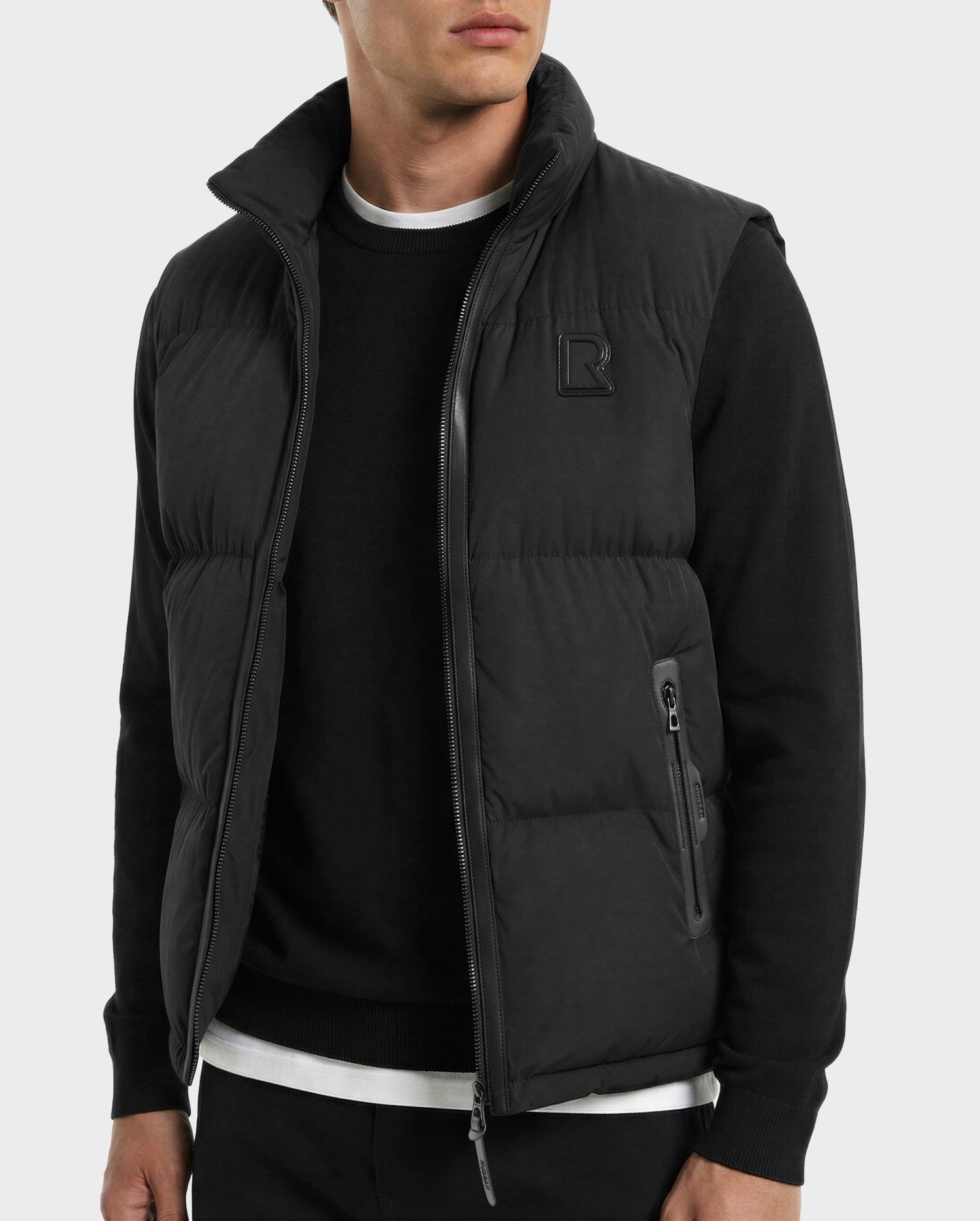A man wears Rudsak's FREDDY - 6125571 Black premium down men's puffer vest over a black sweatshirt with white trim and black pants, standing against a light gray background—perfect for lightweight layering.