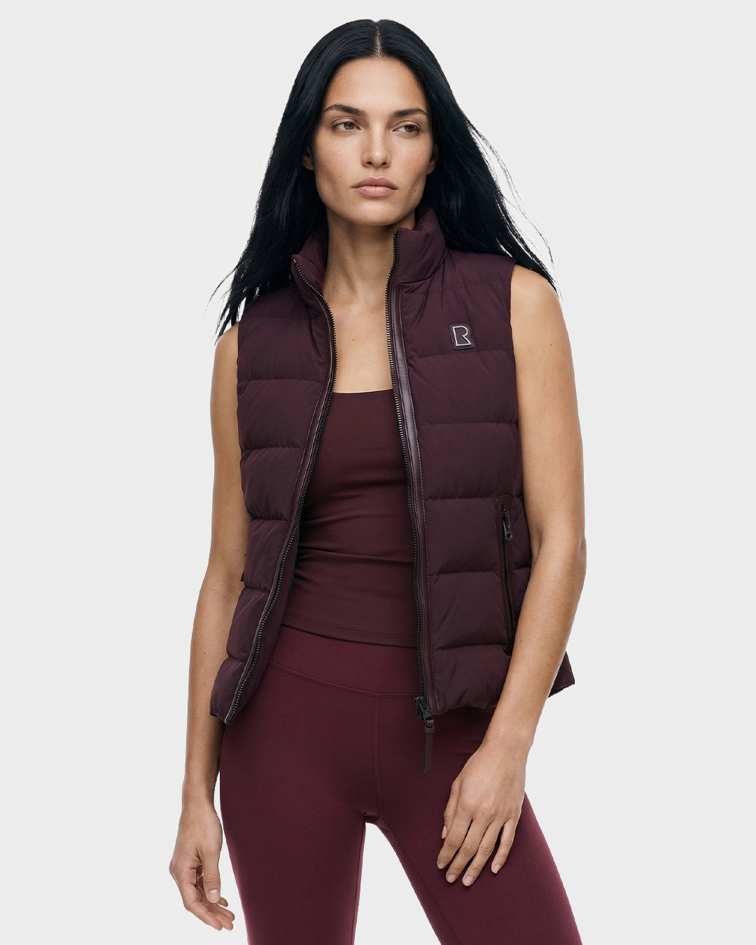 A woman with long black hair wears the Rudsak GABRIELA - 8125014 Bordeaux women’s down puffer vest over a matching tank top and leggings, standing against a plain light gray background.