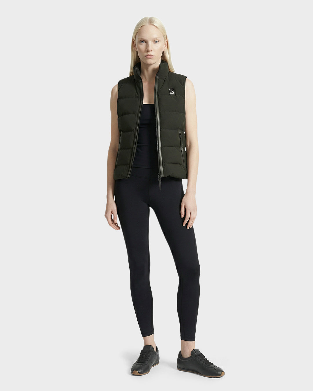 A woman with long blonde hair wears the Rudsak GABRIELA - 8125014 Dark Olive women's lightweight down puffer vest, styled with a black tank top, leggings, and sneakers, standing against a plain light gray background.