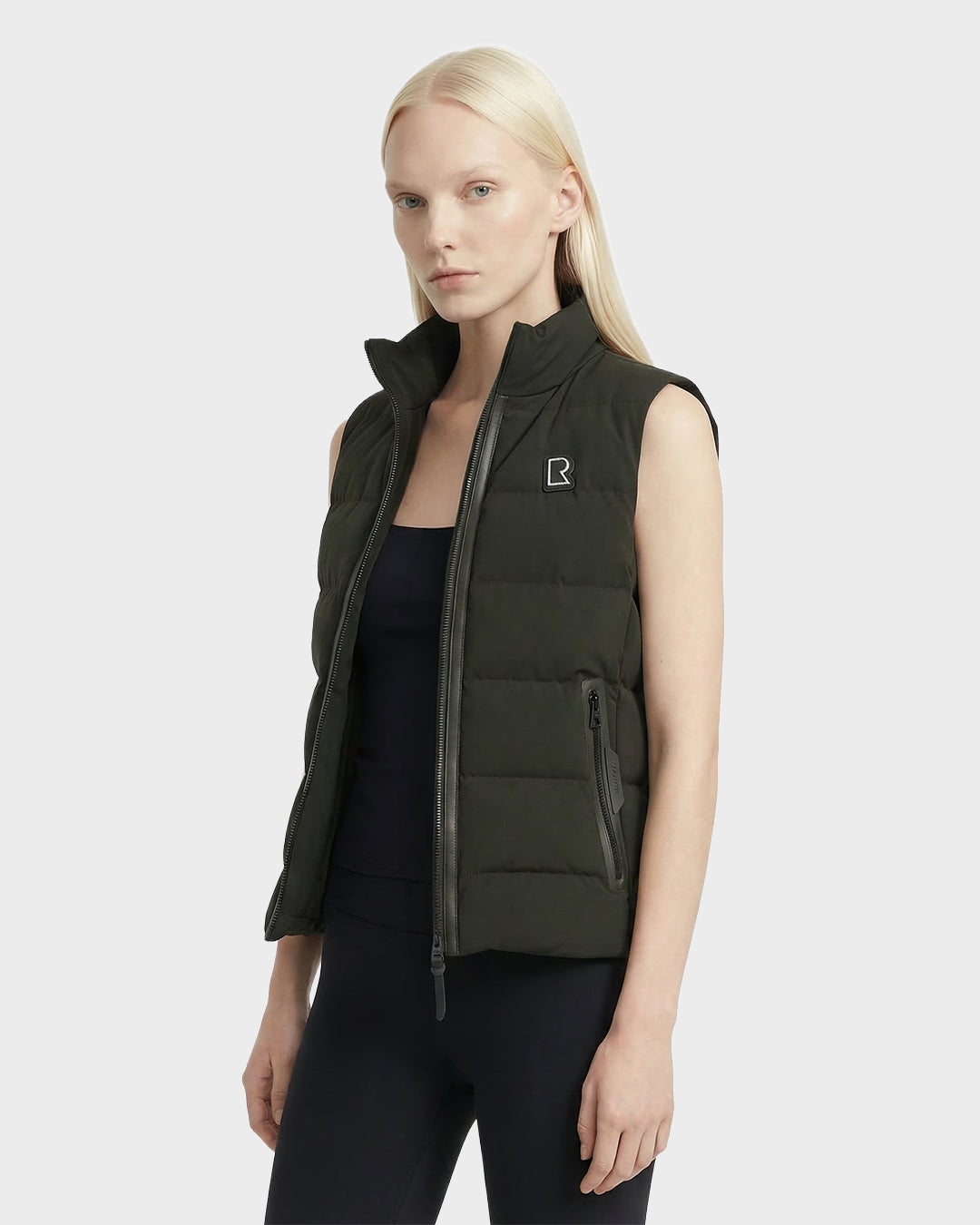 A woman with straight blonde hair wears the Rudsak GABRIELA - 8125014 Dark Olive lightweight down puffer vest with vegan leather trims and a small left-chest logo over a black top and leggings, standing against a plain light background.