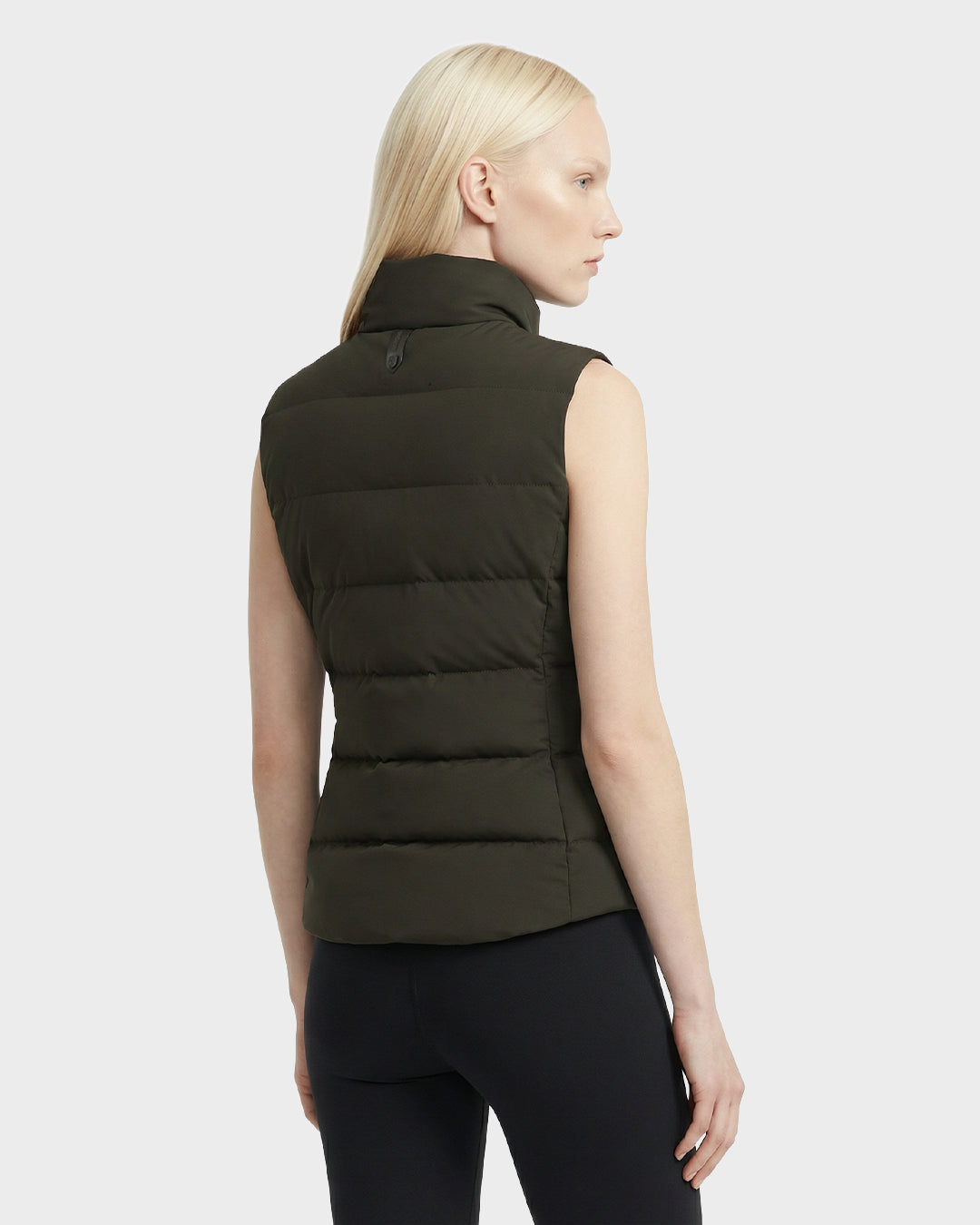 A person with straight light blonde hair wears the Rudsak GABRIELA - 8125014 Dark Olive women’s lightweight down puffer vest with vegan leather trims and black pants, standing angled away against a plain light gray background.