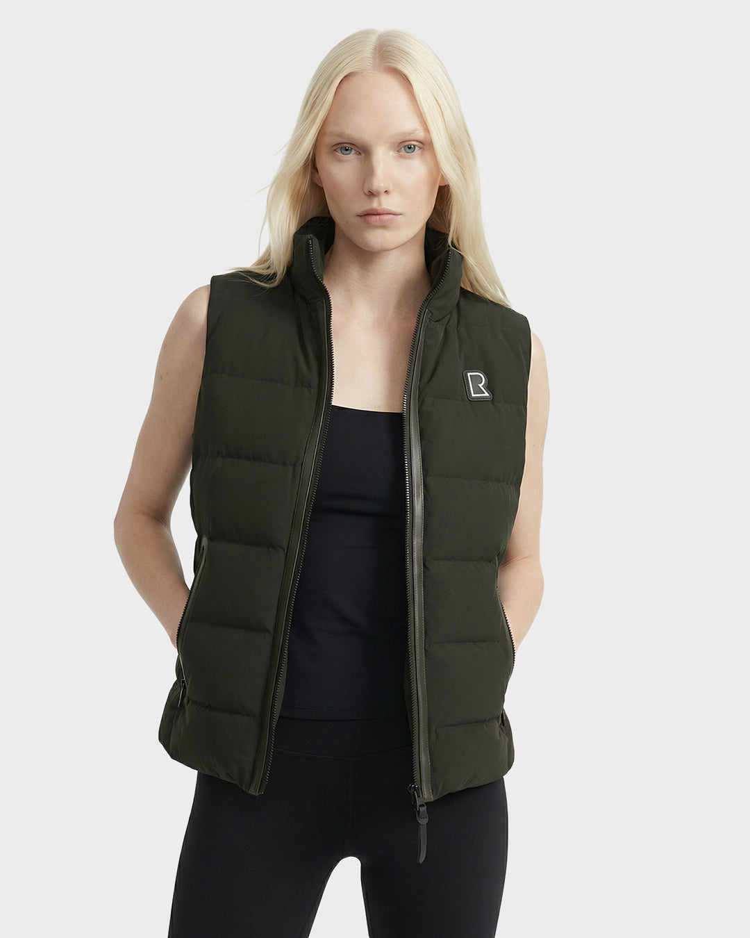 A woman with long blonde hair wears the Rudsak GABRIELA - 8125014 Dark Olive women’s lightweight down puffer vest with vegan leather trims over a black outfit, standing hands in pockets against a plain light gray background.